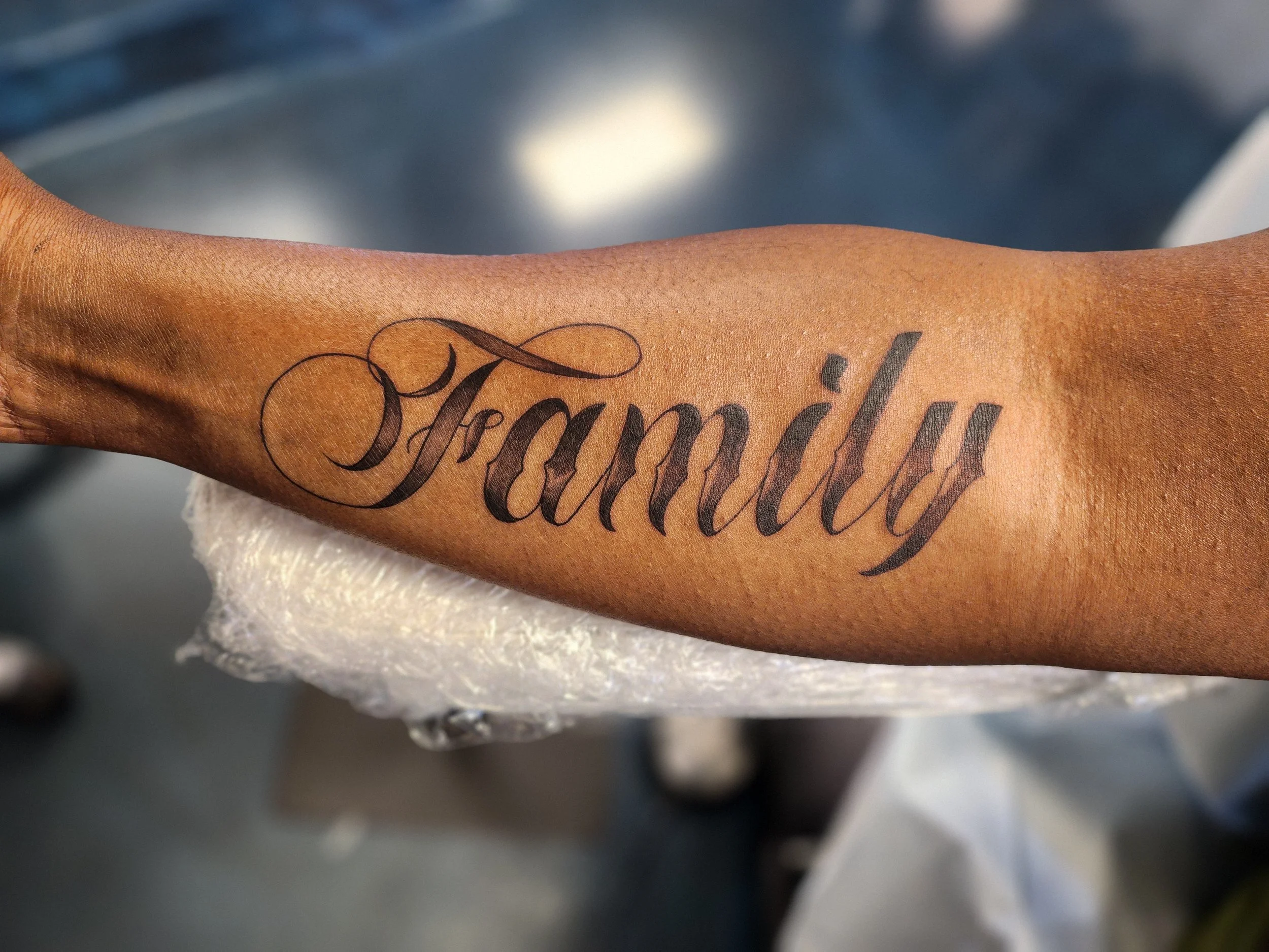 Tattoo of the word 'Family' in elaborate cursive on a person's forearm.