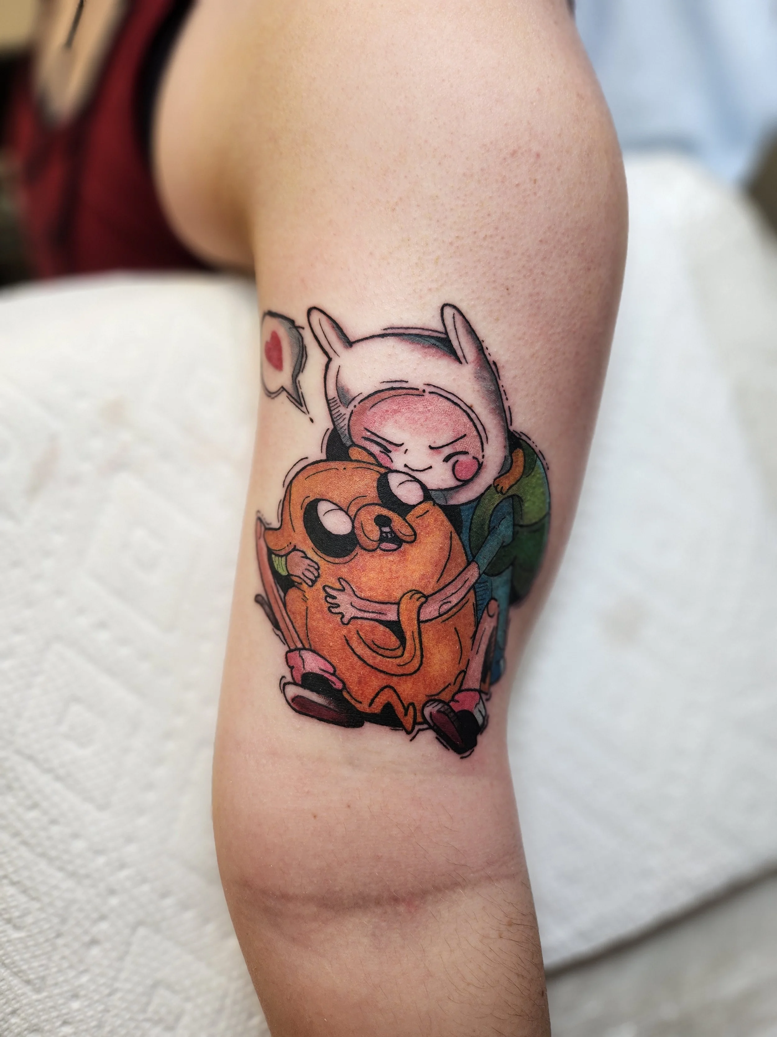 Tattoo of a cartoon girl hugging a round orange creature on a person's upper arm.