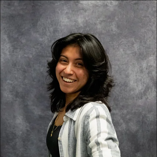 A young woman with shoulder-length black hair smiling and wearing a striped blazer over a dark top, standing against a gray textured background.