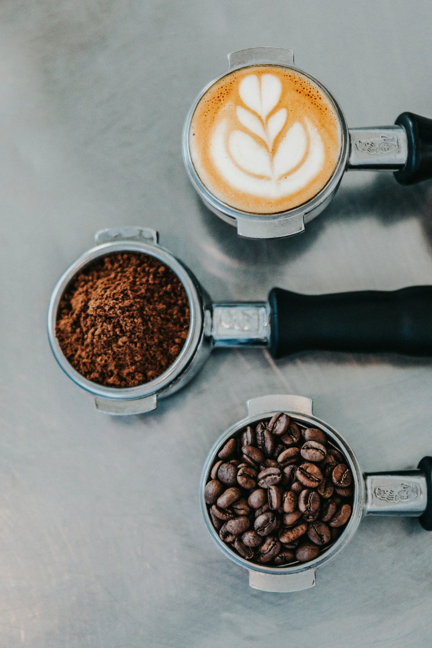 A top-down view of a coffee-making setup with a glass pitcher of latte art, a scoop of ground coffee, and a scoop of whole coffee beans on a gray surface.