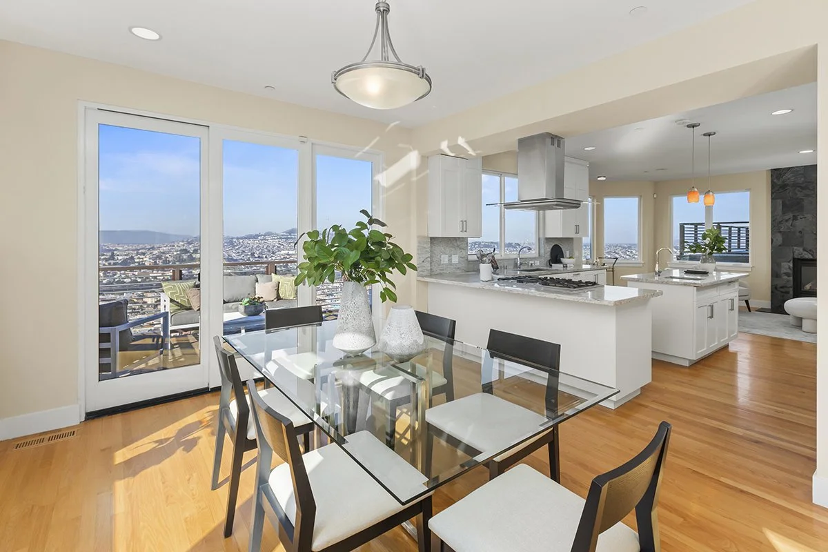 Dining area with glass-top table, black chairs, and a large plant in white vases, overlooking a balcony with outdoor seating and a cityscape view through large windows and sliding doors.
