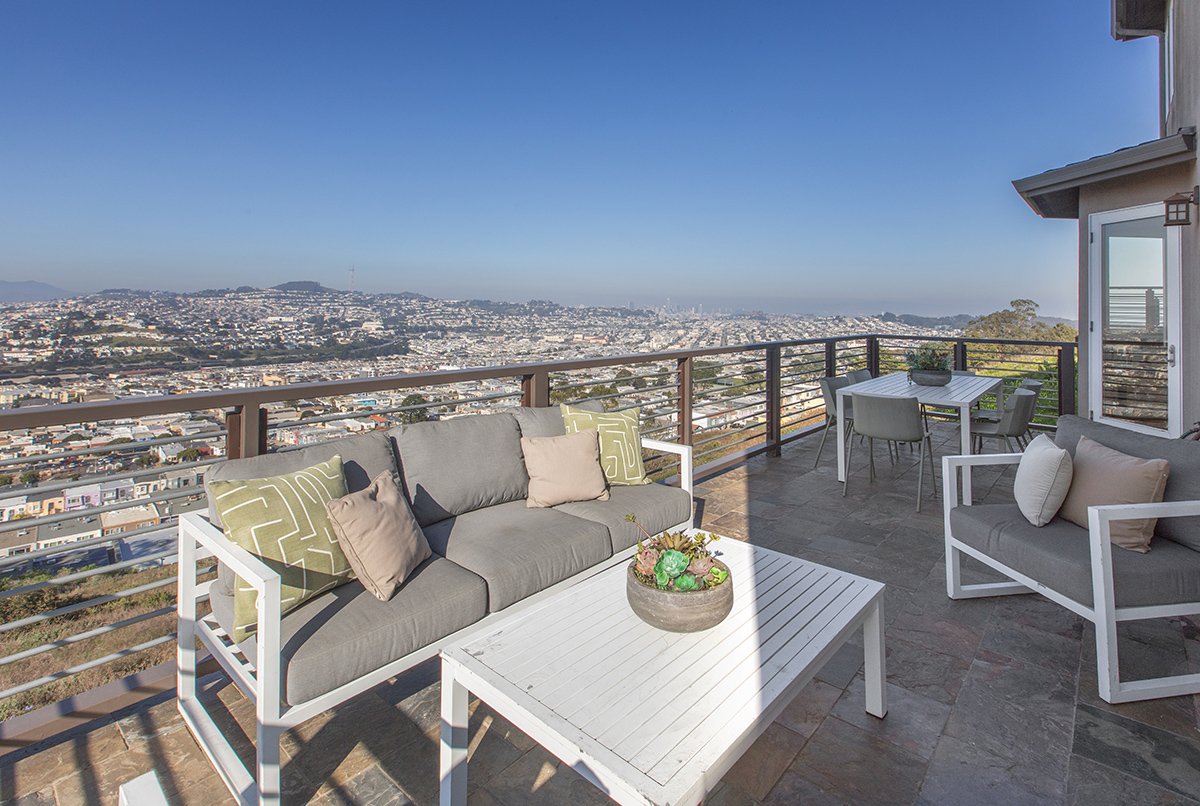 Outdoor balcony with gray sofa, chairs, and a table, overlooking a cityscape under a clear blue sky.