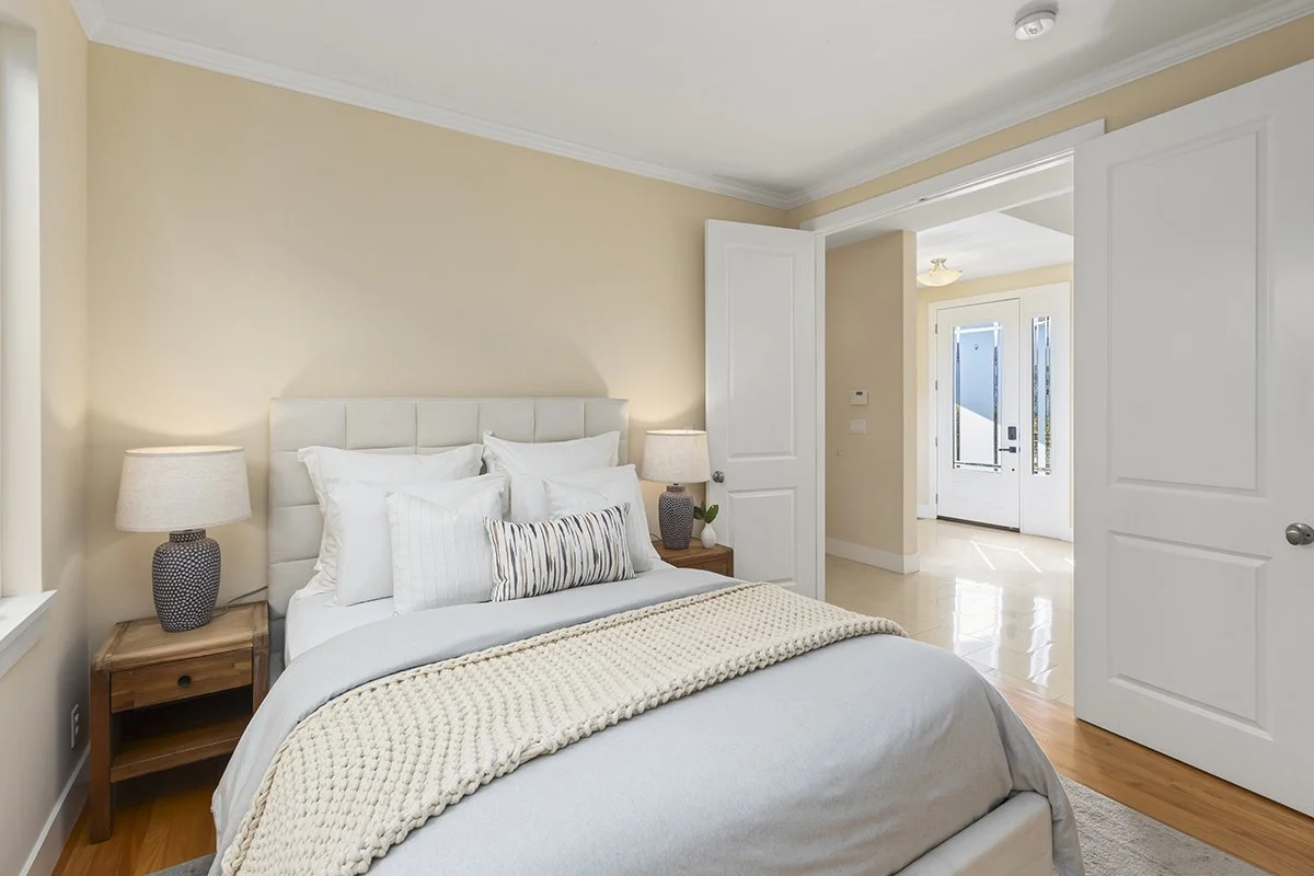 A bedroom with a bed dressed in white linens and decorative pillows, flanked by two wooden nightstands with lamps. An open door leads to a hallway or entryway.