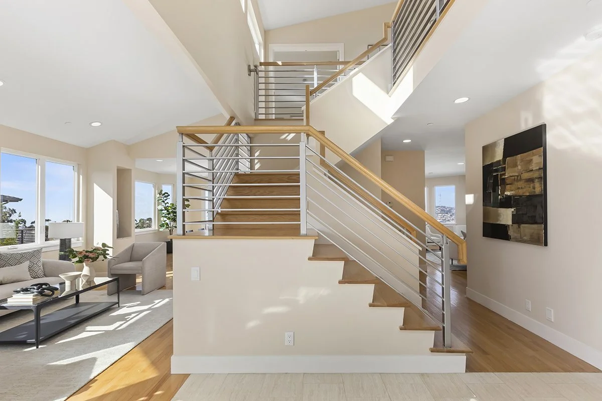 Modern living room with wooden staircase, brass and wood railing, sunlight streaming in through large windows, minimalistic furniture, and a decorative wall art piece.