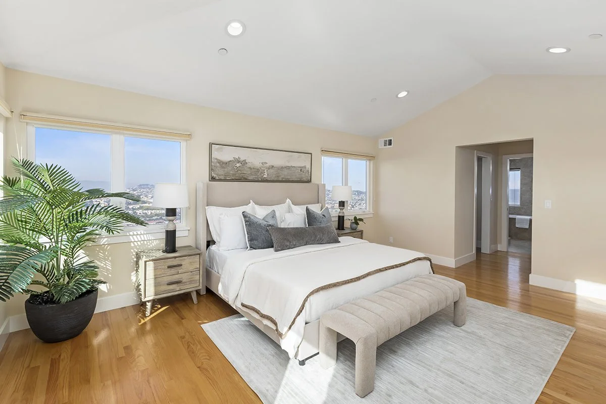 A modern bedroom with a large bed, gray and white bedding, upholstered bench at the foot of the bed, two nightstands with lamps, a tall green plant in a black pot, and windows showing a cityscape view.