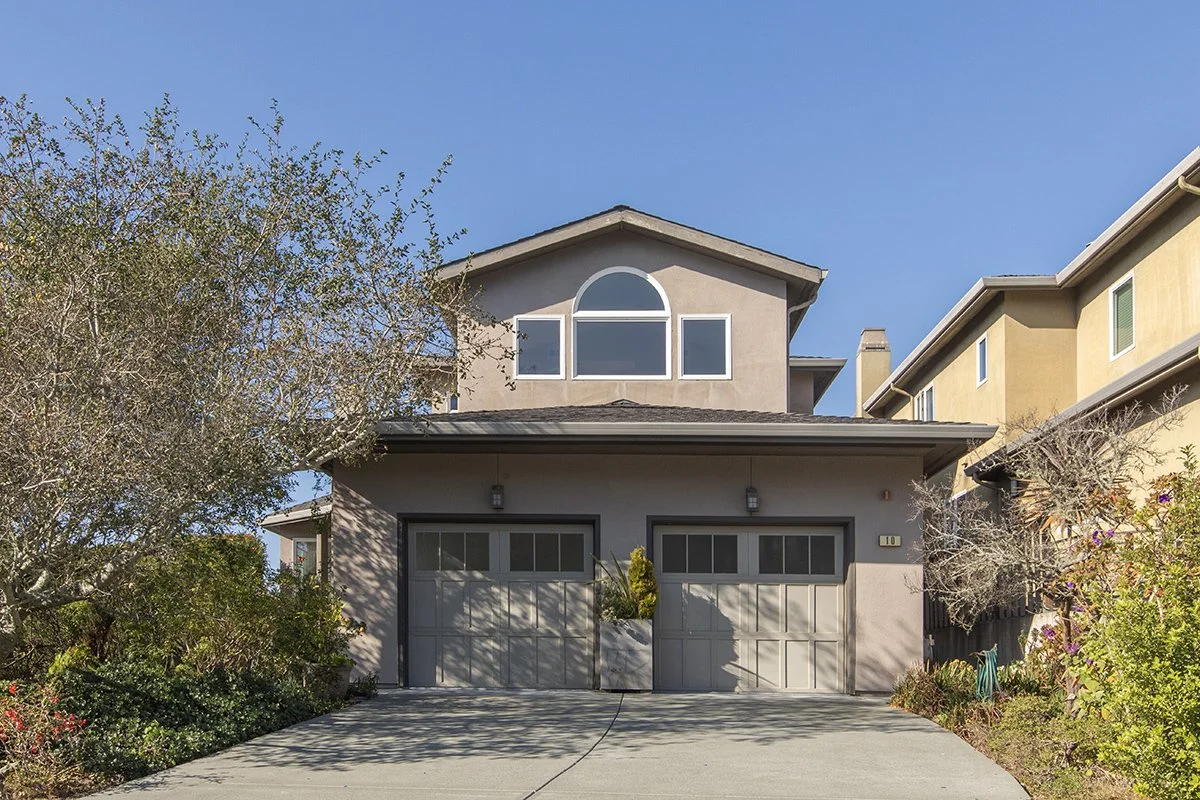 A two-story house with a garage, beige and gray exterior, and a large arched window on the upper floor, with trees and plants in the front yard.