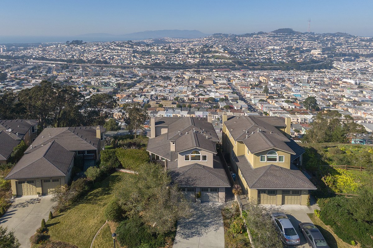 Aerial view of a hillside neighborhood with three houses, surrounded by greenery, overlooking a densely packed cityscape with many buildings and streets in the distance.