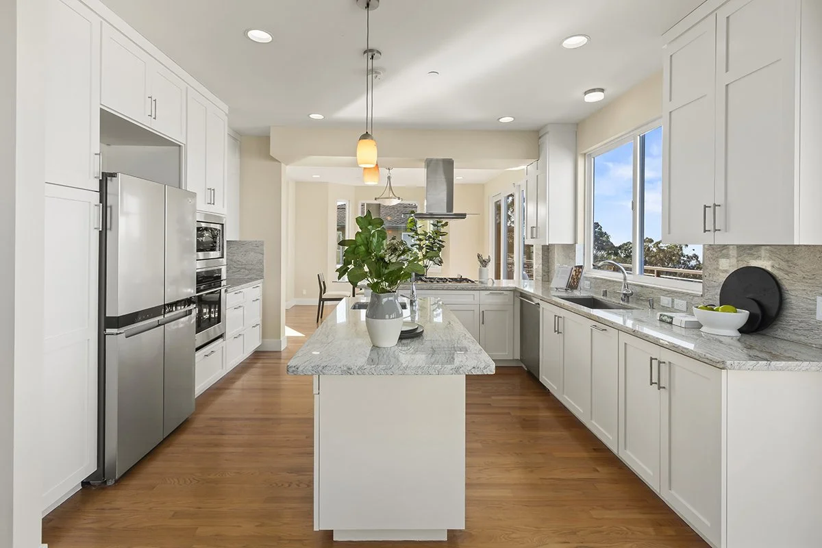 Bright, modern kitchen with white cabinets, granite countertops, stainless steel appliances, and hardwood floors. Large window with a view outside, and a central island with a plant in a white vase.