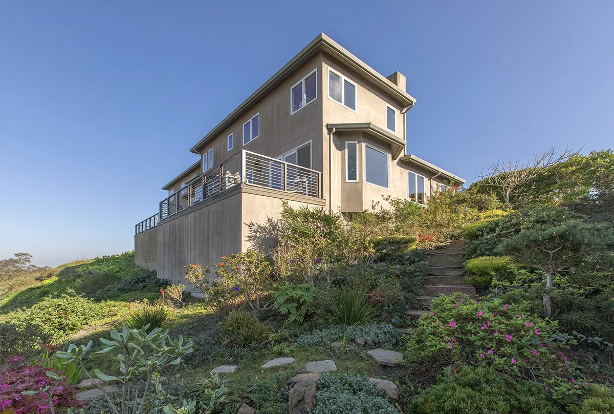 A modern multi-story house on a hill with large windows and a balcony, surrounded by lush greenery and plants.