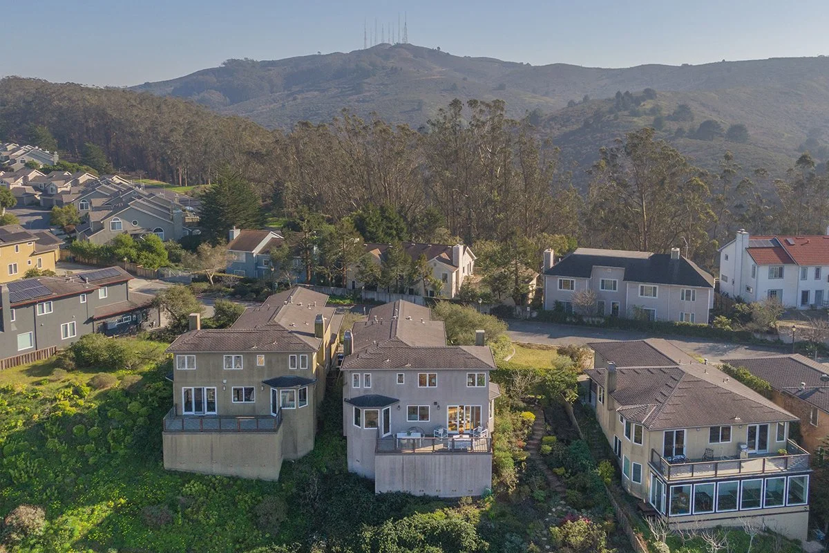 Aerial view of coastal residential neighborhood with multi-story houses, greenery, and mountains in background.