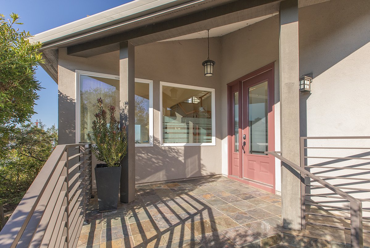 Front porch of a modern house with a red front door, large windows, potted plant, and slate tile flooring under a covered roof, with shadows cast on the floor.