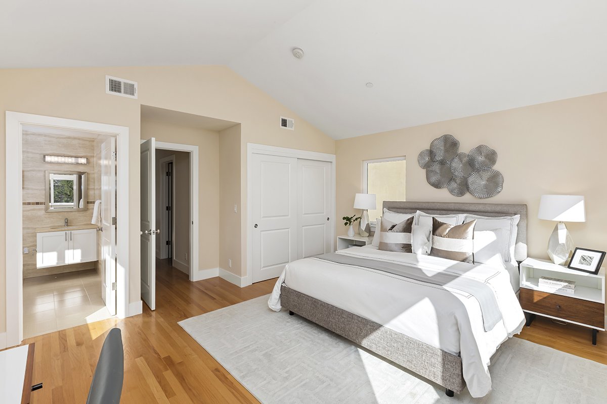 Bright bedroom with a bed, nightstands, lamps, and wall art, alongside a view into an en-suite bathroom.