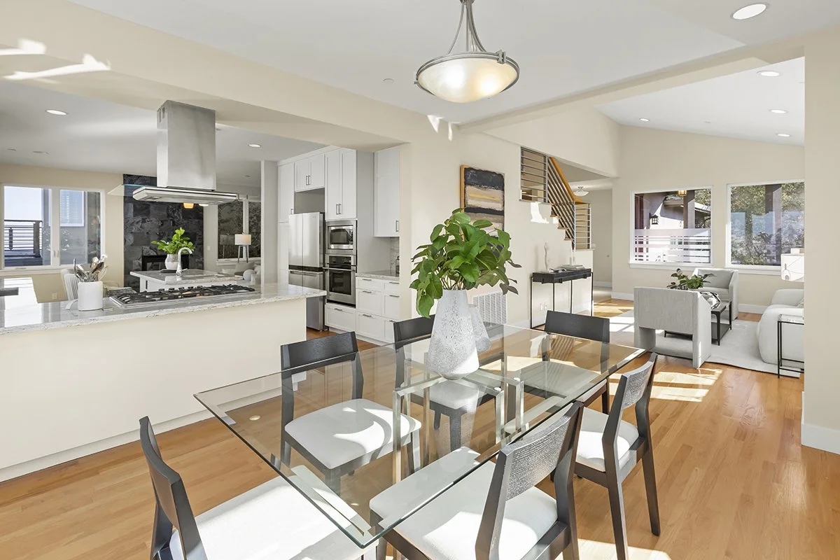 Open-concept kitchen and dining area with a glass dining table, black chairs, white cabinetry, stainless steel appliances, and a living room with sofas and large windows.