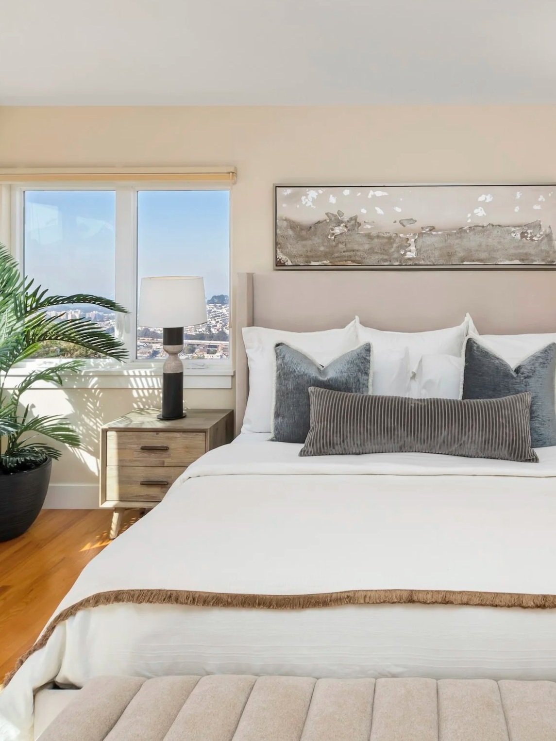 Bright living room with large windows overlooking a cityscape, featuring a staircase with metal railing, a dining area with a table and chairs, and contemporary furniture with plants and decor.