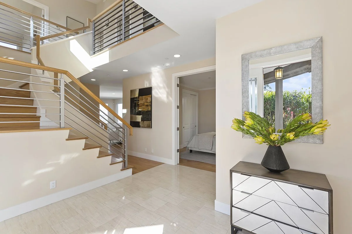 Interior view of a modern, bright house with stairs, a mirror with a flower arrangement, and a bedroom visible through an open door.