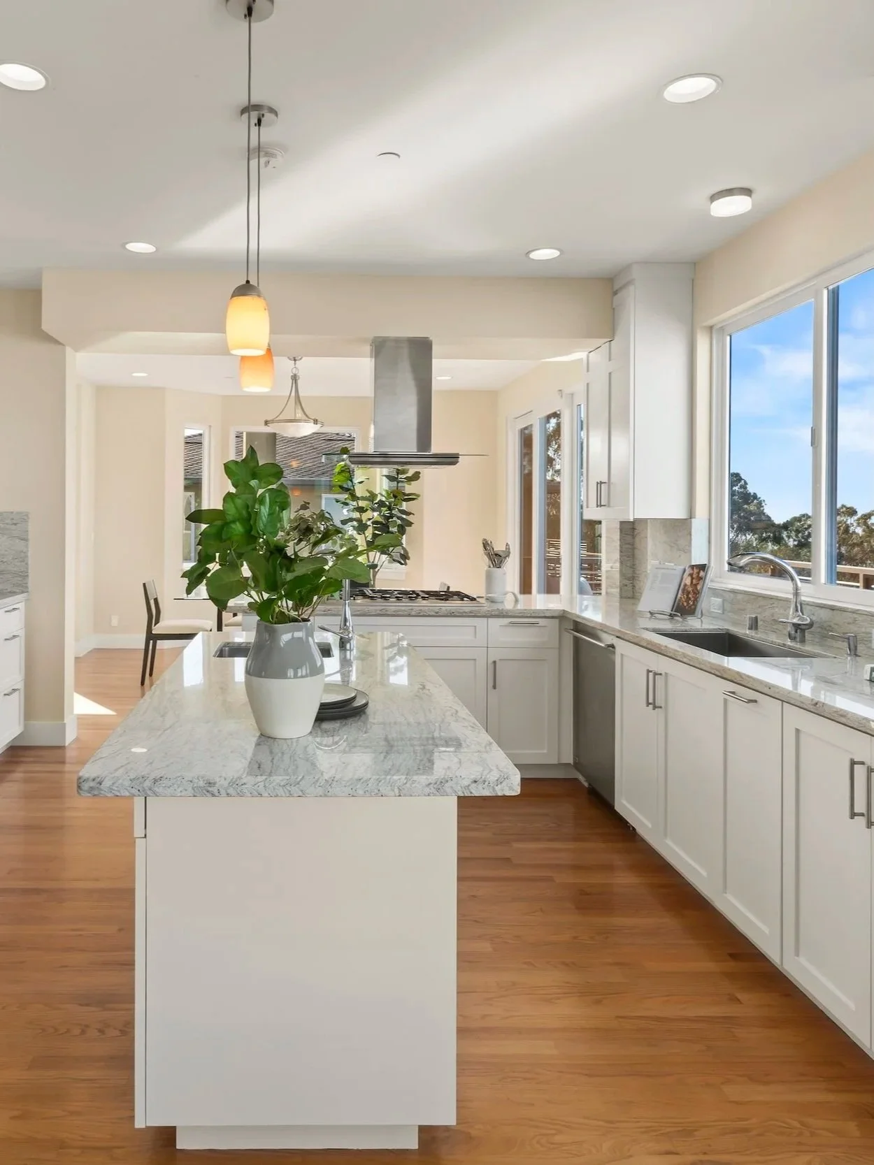 Modern kitchen with white cabinets, granite countertops, and a kitchen island with a potted plant, pendant lighting, and large windows showing blue sky outside.