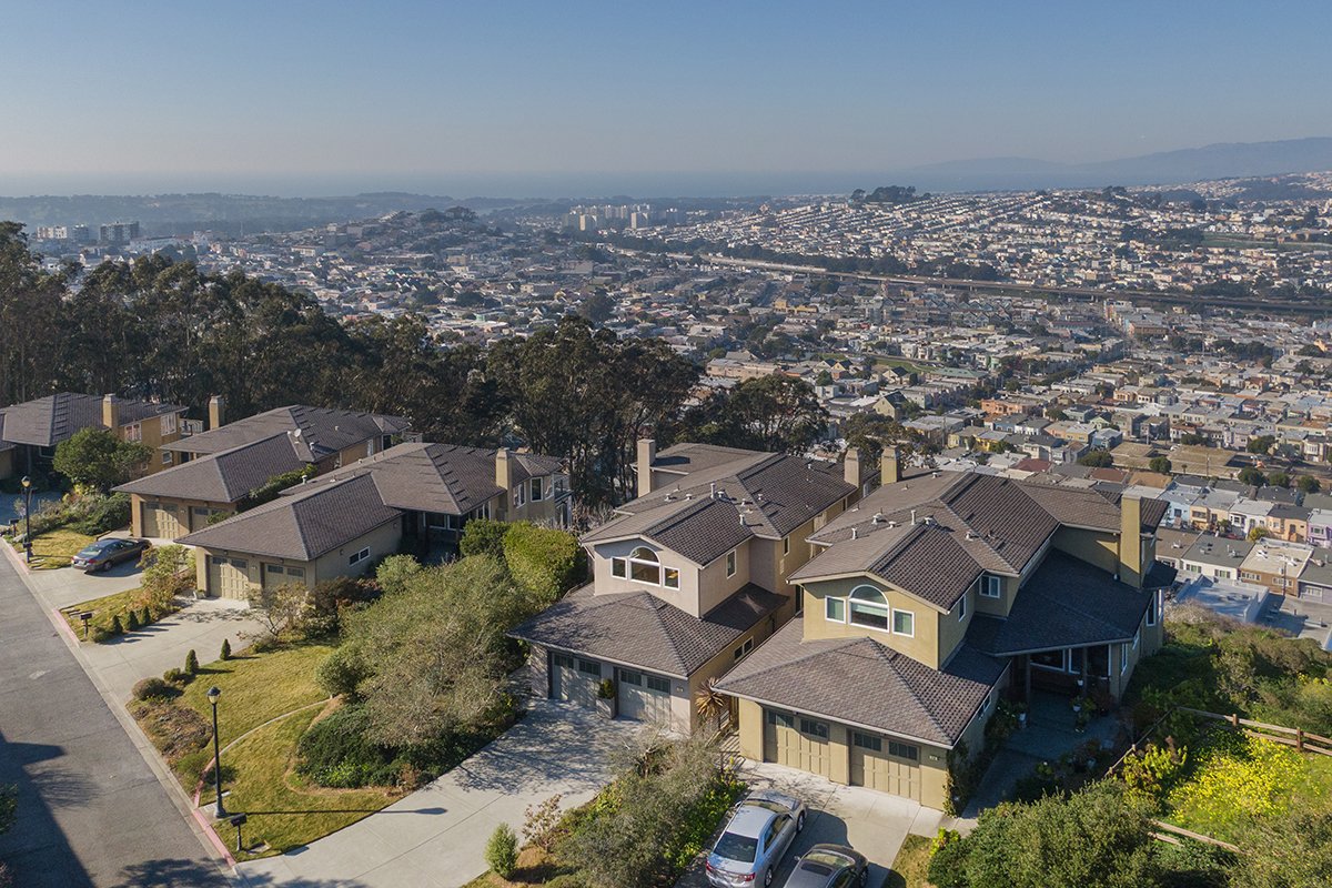 Aerial view of a suburban neighborhood with multiple houses, some with garages, surrounded by trees, with a cityscape in the distance and hills on the horizon under a clear blue sky.