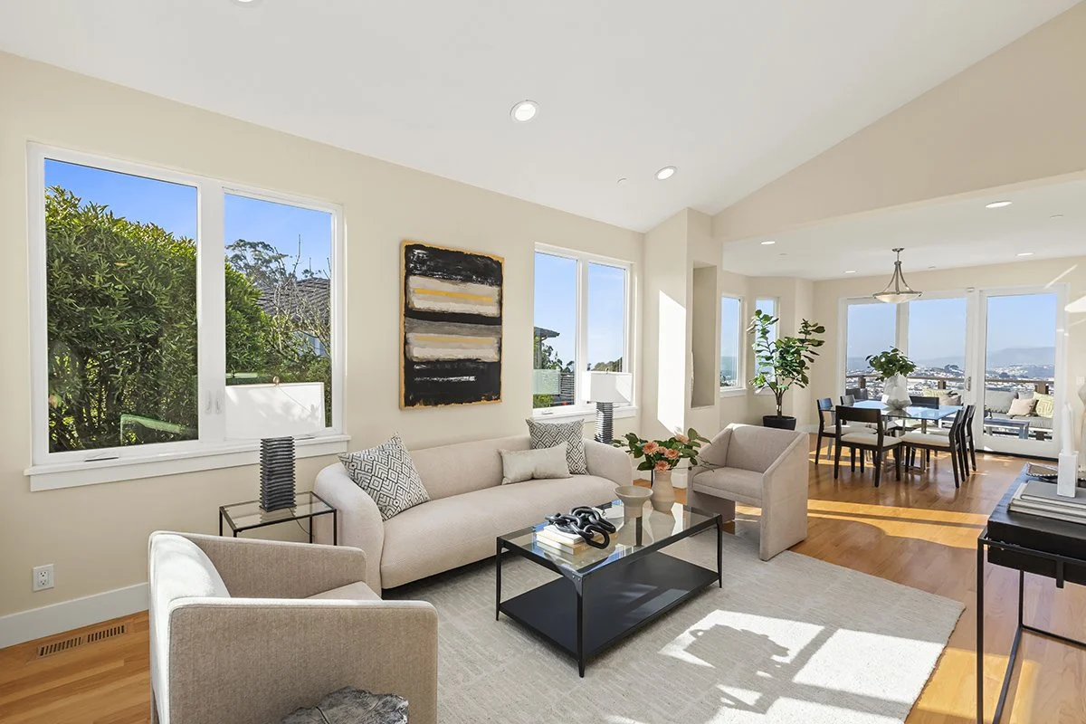 Bright living room with beige sofas, a glass coffee table, and large windows showing blue sky and greenery outside.