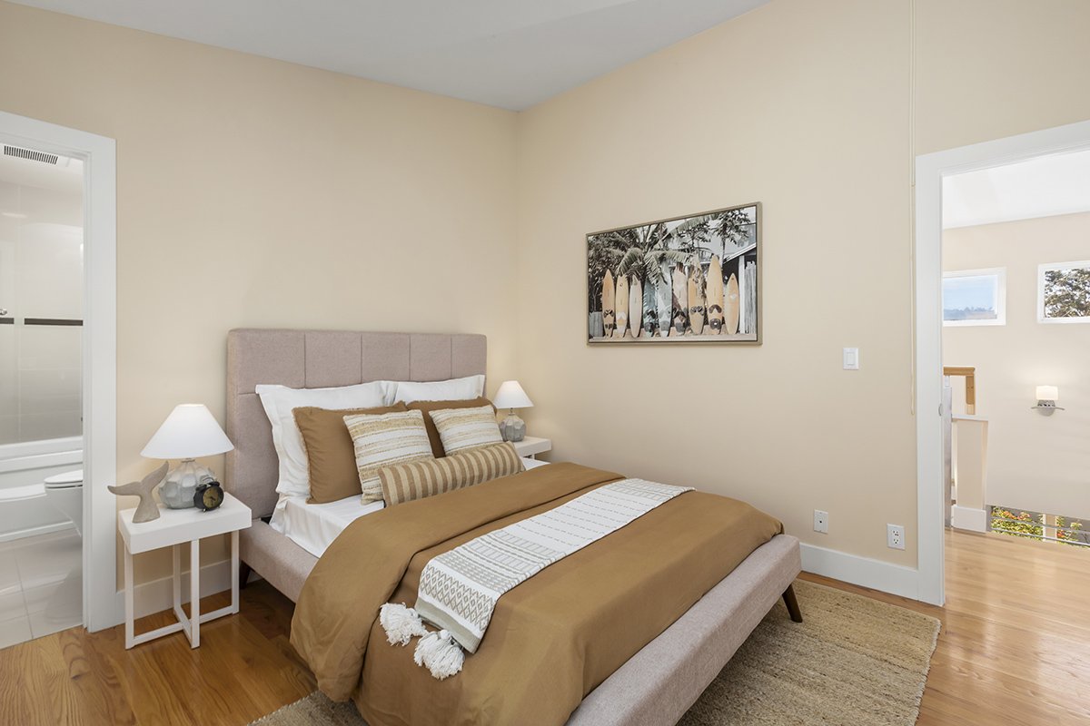 A cozy bedroom with a beige upholstered bed, white and brown pillows, brown duvet, and a decorative throw blanket at the foot of the bed. Two white nightstands with lamps, a decorative starfish, and a clock sit beside the bed. A framed beach-themed p