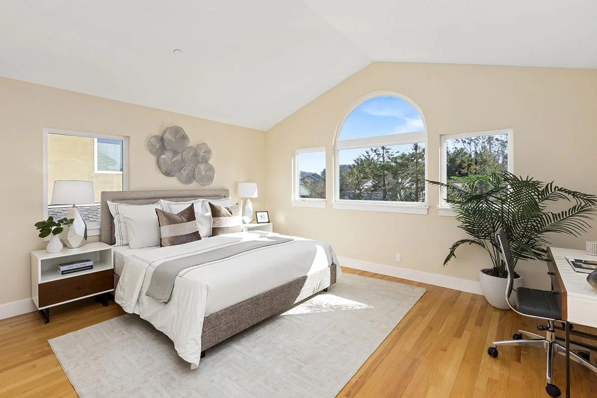 Bright bedroom with a large bed, white bedding, and multiple pillows, beside two nightstands with lamps and a plant, with large windows showing trees and blue sky outside, and a desk with a chair and a large potted plant.
