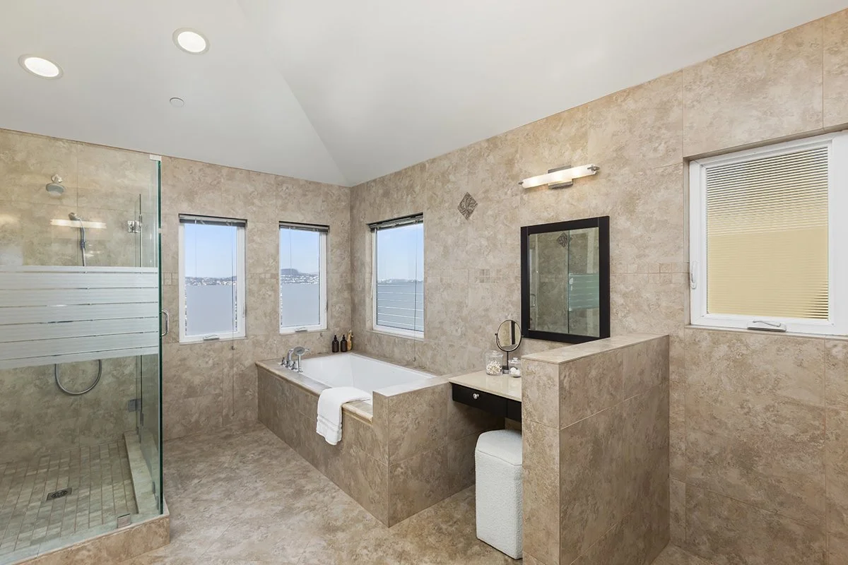 Modern bathroom with a whirlpool tub, glass-enclosed shower, three windows with a view of water, beige marble walls and floor, and a vanity with mirror and light fixture.