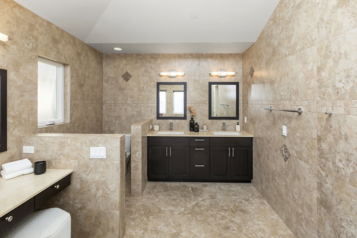 A bathroom with beige tiled walls and floor, featuring a double vanity with two mirrors, two sinks, and modern lighting fixtures above. There is a small window on the left, a towel rack on the right wall, and a partial wall separating the toilet area