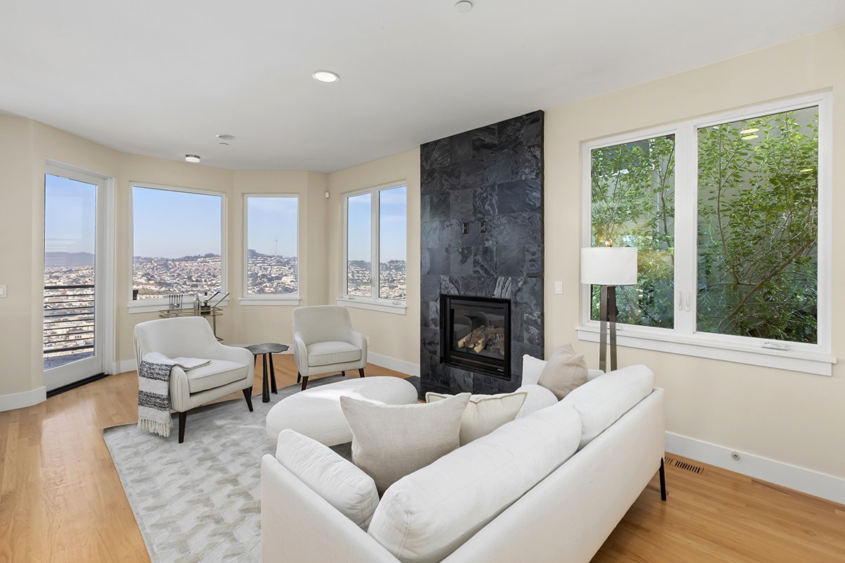Bright living room with white sofa, armchairs, black marble fireplace, large windows showing city views, and greenery outside.