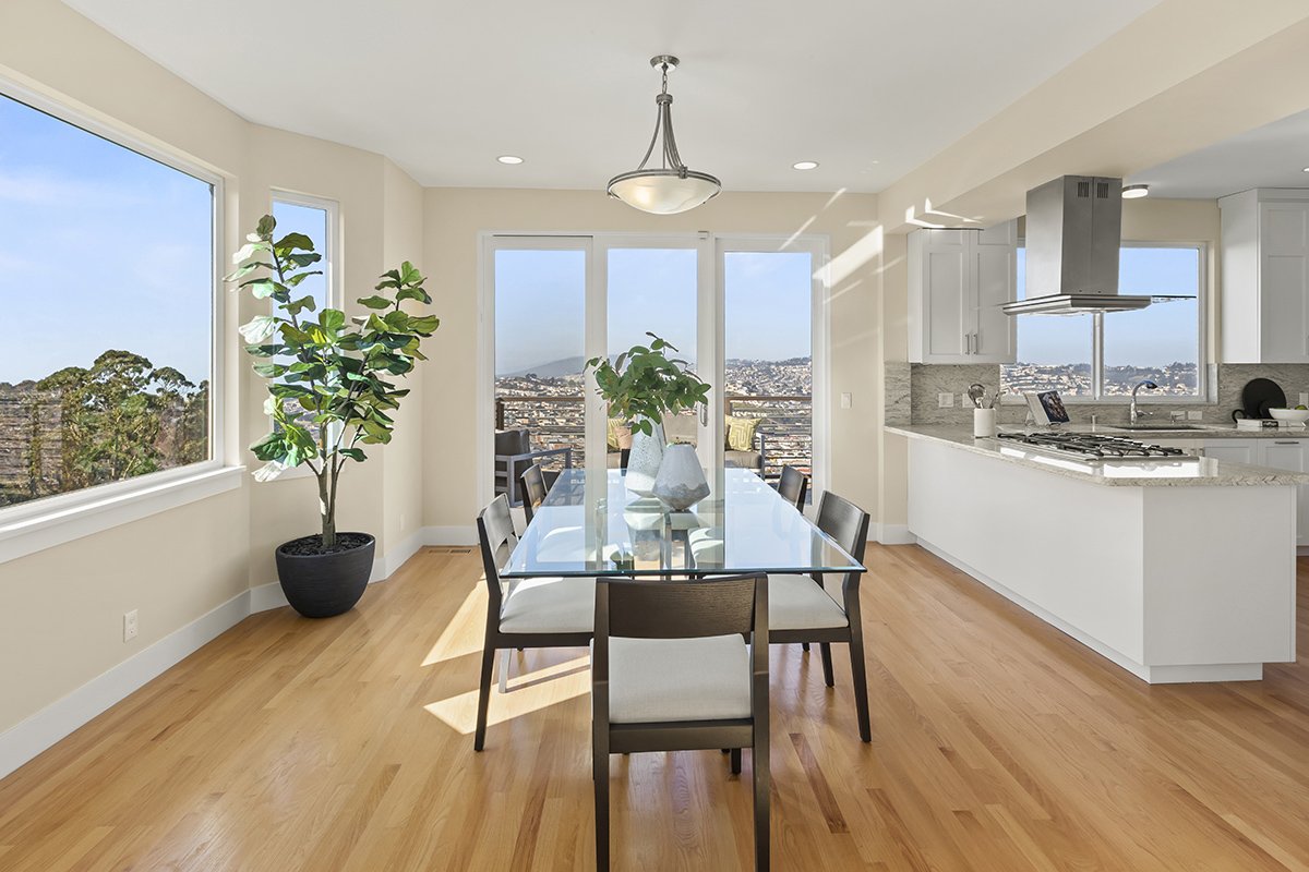 Modern dining area with a glass table, eight chairs, large potted plants, bright windows, and a view of a cityscape.