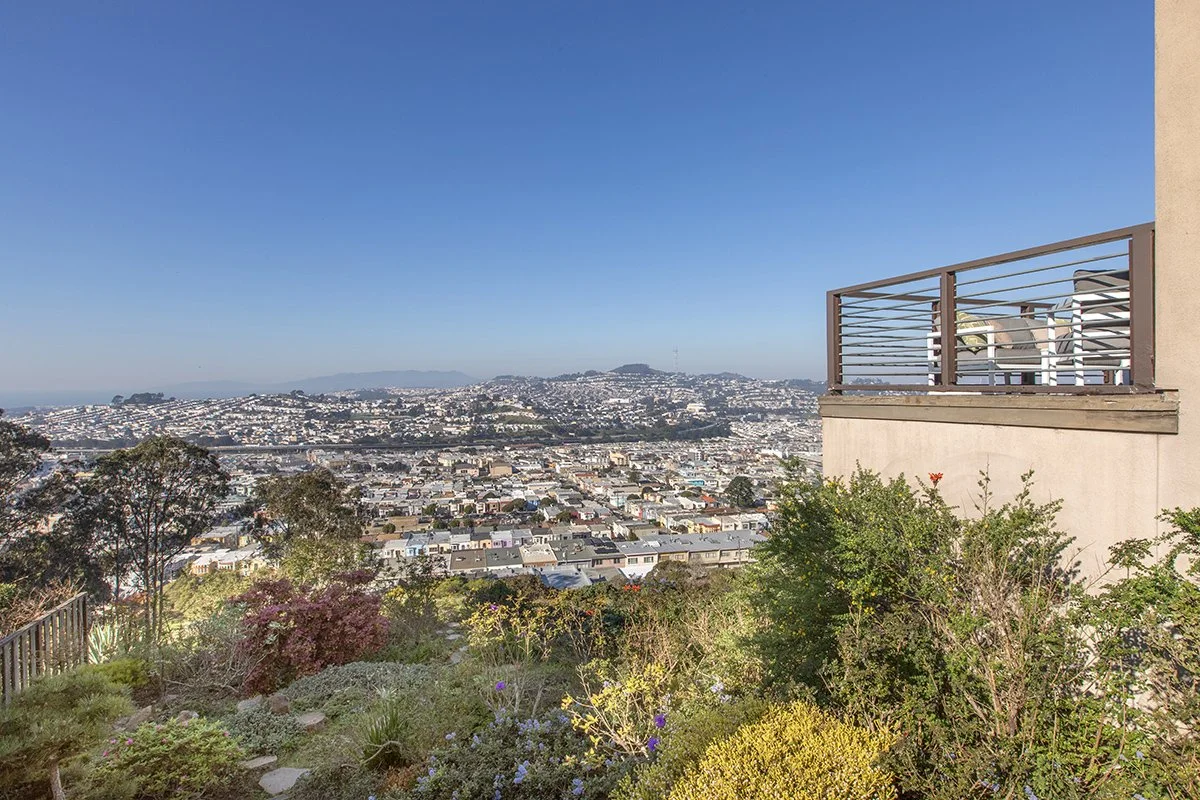 View of a hilly cityscape with numerous white and light-colored buildings, a clear blue sky, and a small balcony with chairs on the right side of the image. Foreground includes a garden with green bushes and flowering plants.