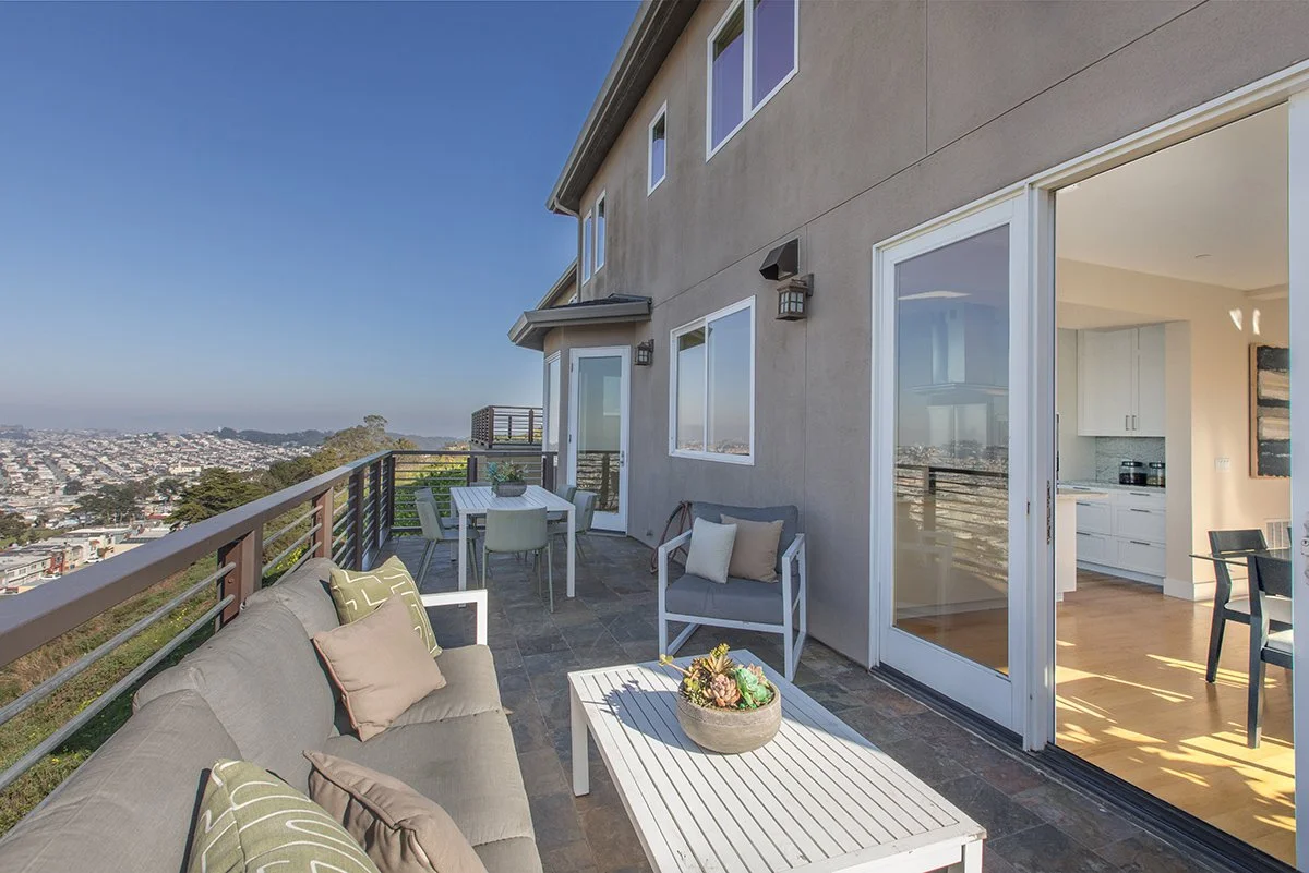 Balcony with outdoor seating and a view of a cityscape, adjacent to an open kitchen.