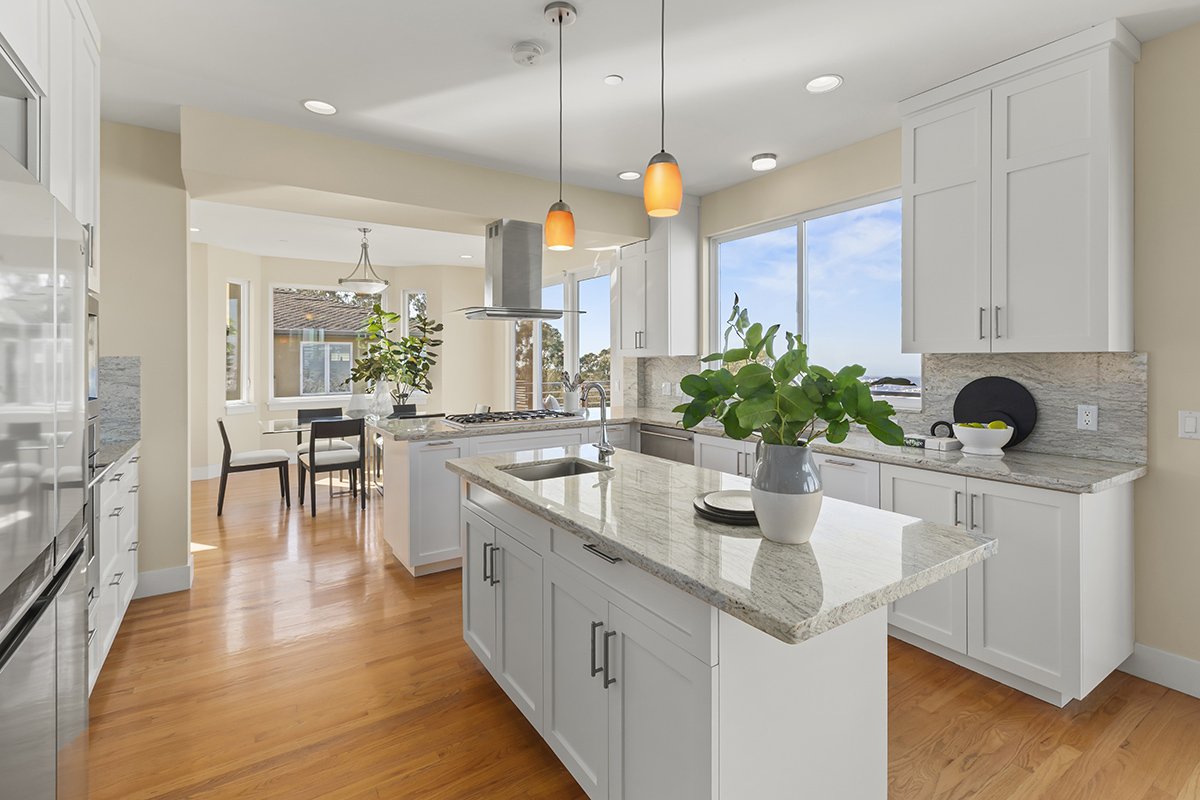 Modern kitchen with white cabinets, granite countertops, hardwood floors, pendant lights, and large windows with a view of the sky.
