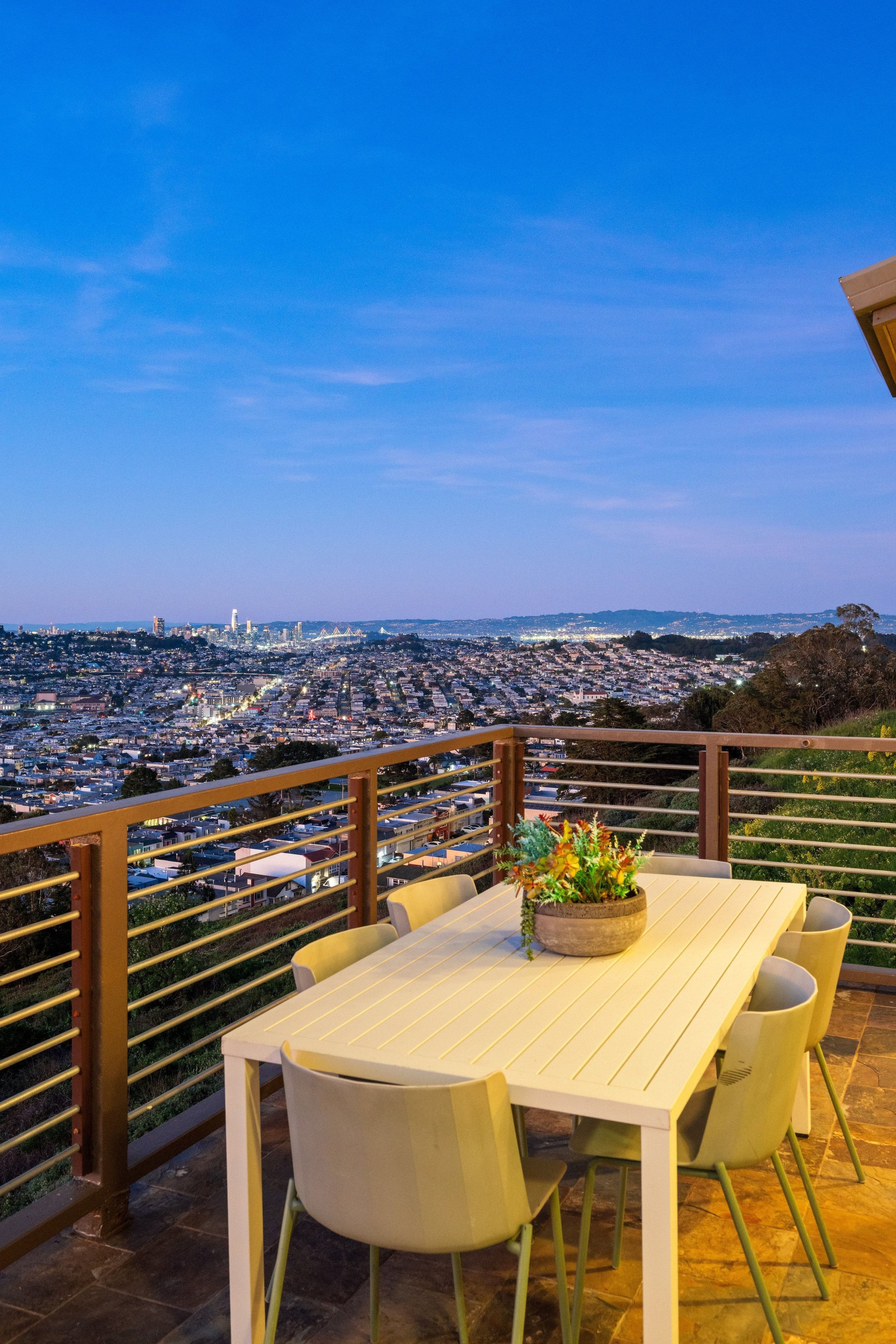 Outdoor rooftop balcony with gray sofa, various pillows, white coffee table with plant, overlooking cityscape and hills under clear blue sky.