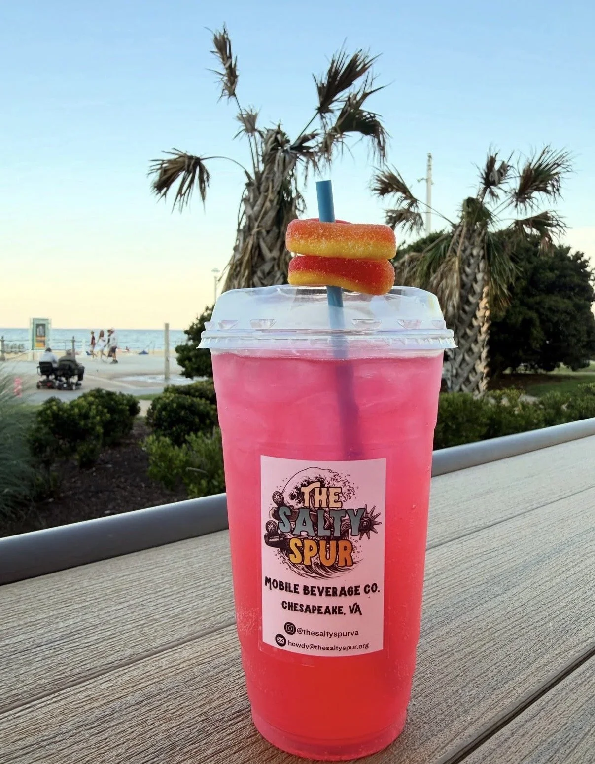 Pink beverage in a clear plastic cup with a straw and two peach rings on top, outdoors near the beach with palm trees and people in the background.