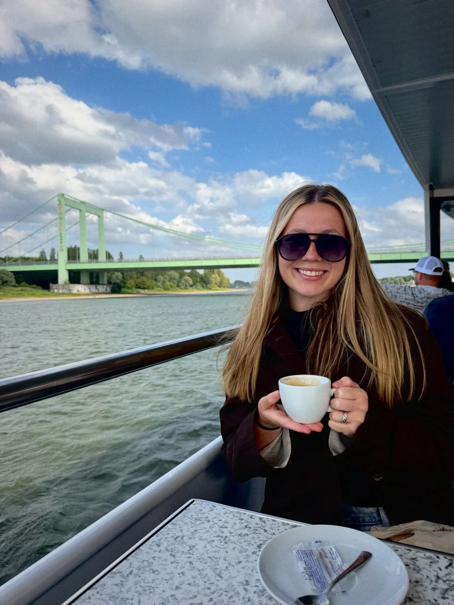 A woman with long blonde hair wearing dark sunglasses is smiling and holding a white mug with a hot beverage. She is on a boat or ferry with water and a bridge in the background, and there is a white plate with a spoon and a small packet on the table in front of her.