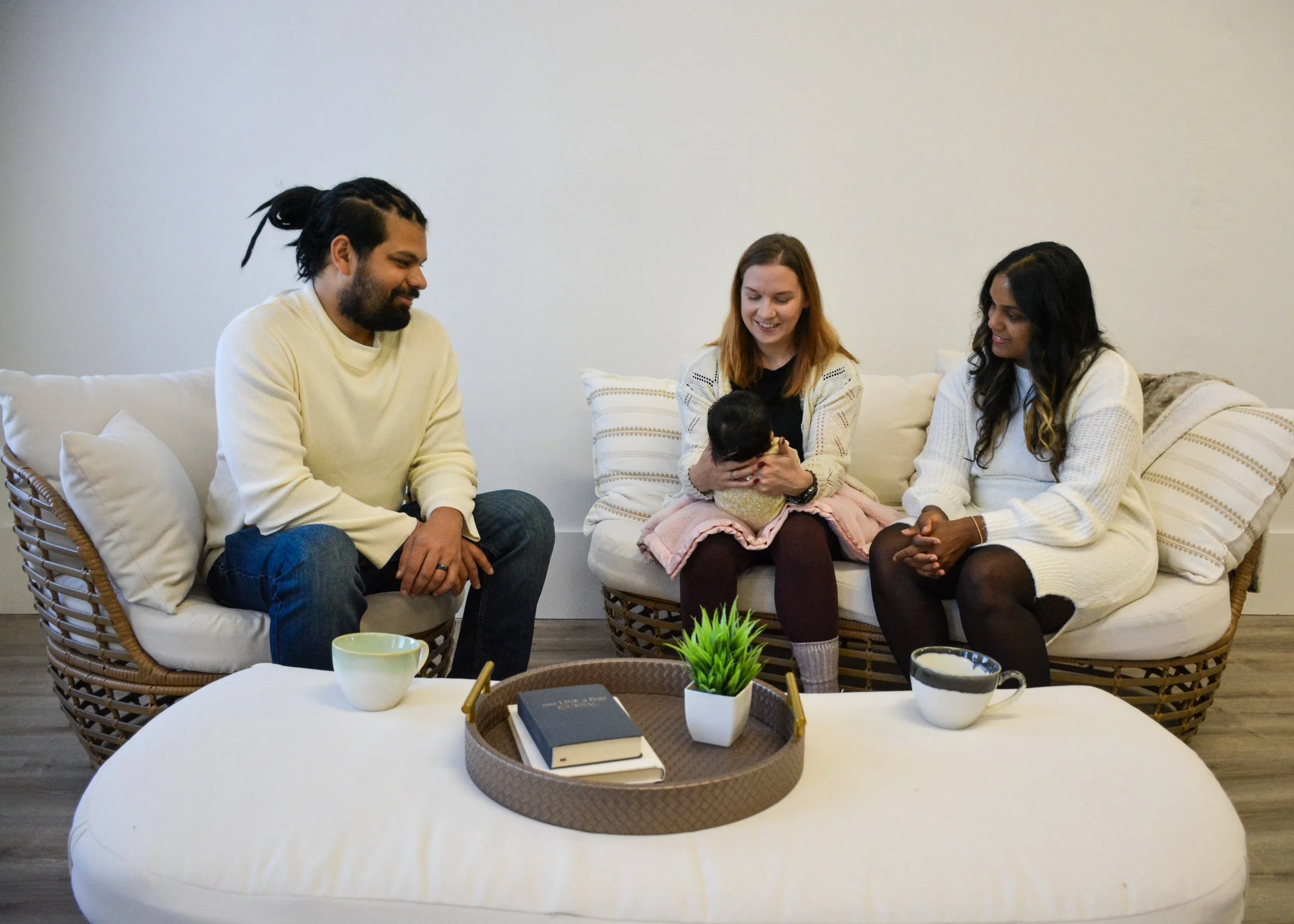 Two women and one man sitting on a white cushioned sofa, with a woman holding a baby, in a cozy living room with a coffee table and decorative pillows.