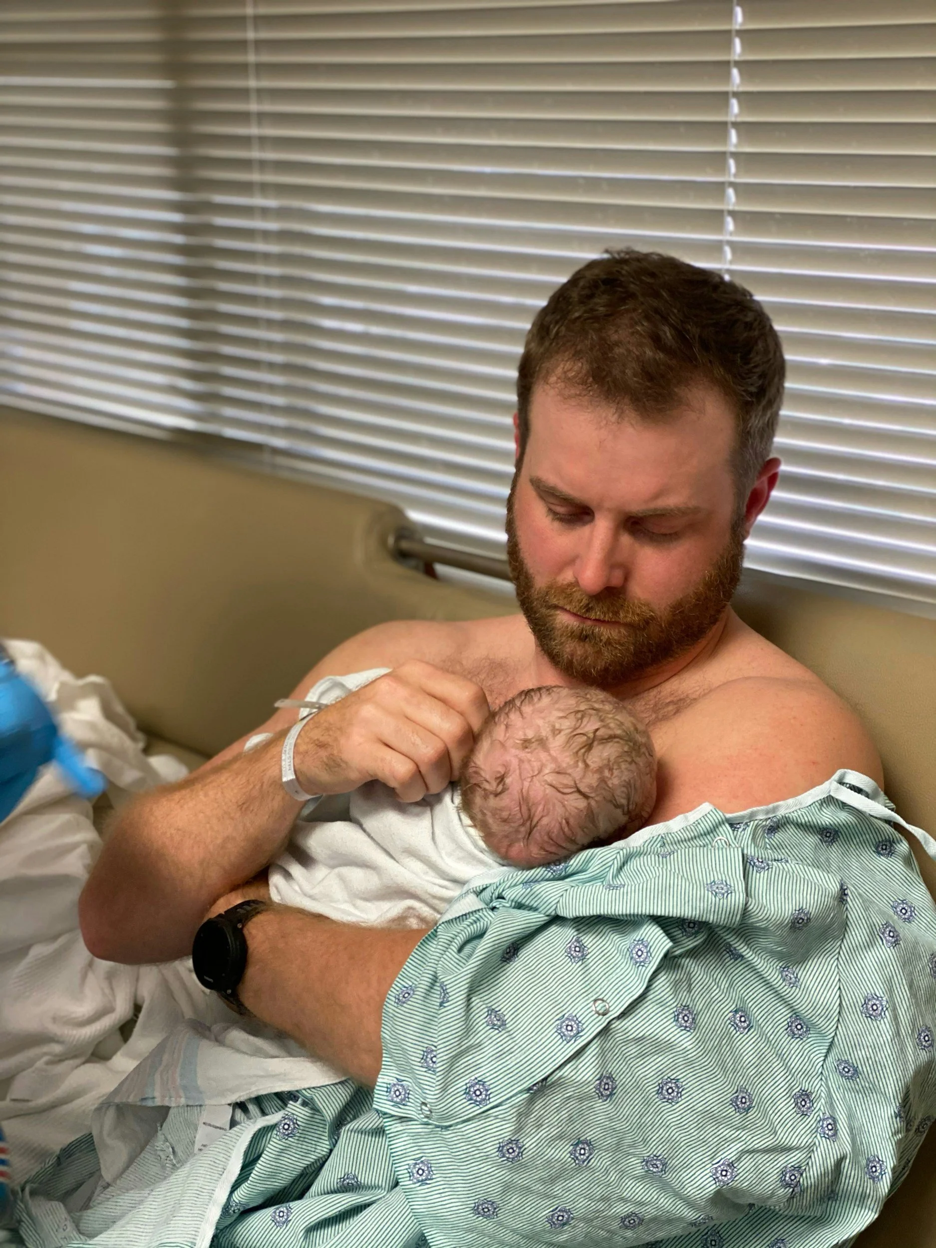 A man in a hospital gown is holding a newborn baby, looking down at the baby with a tender expression. The man is seated on a hospital bed with blinds in the background.
