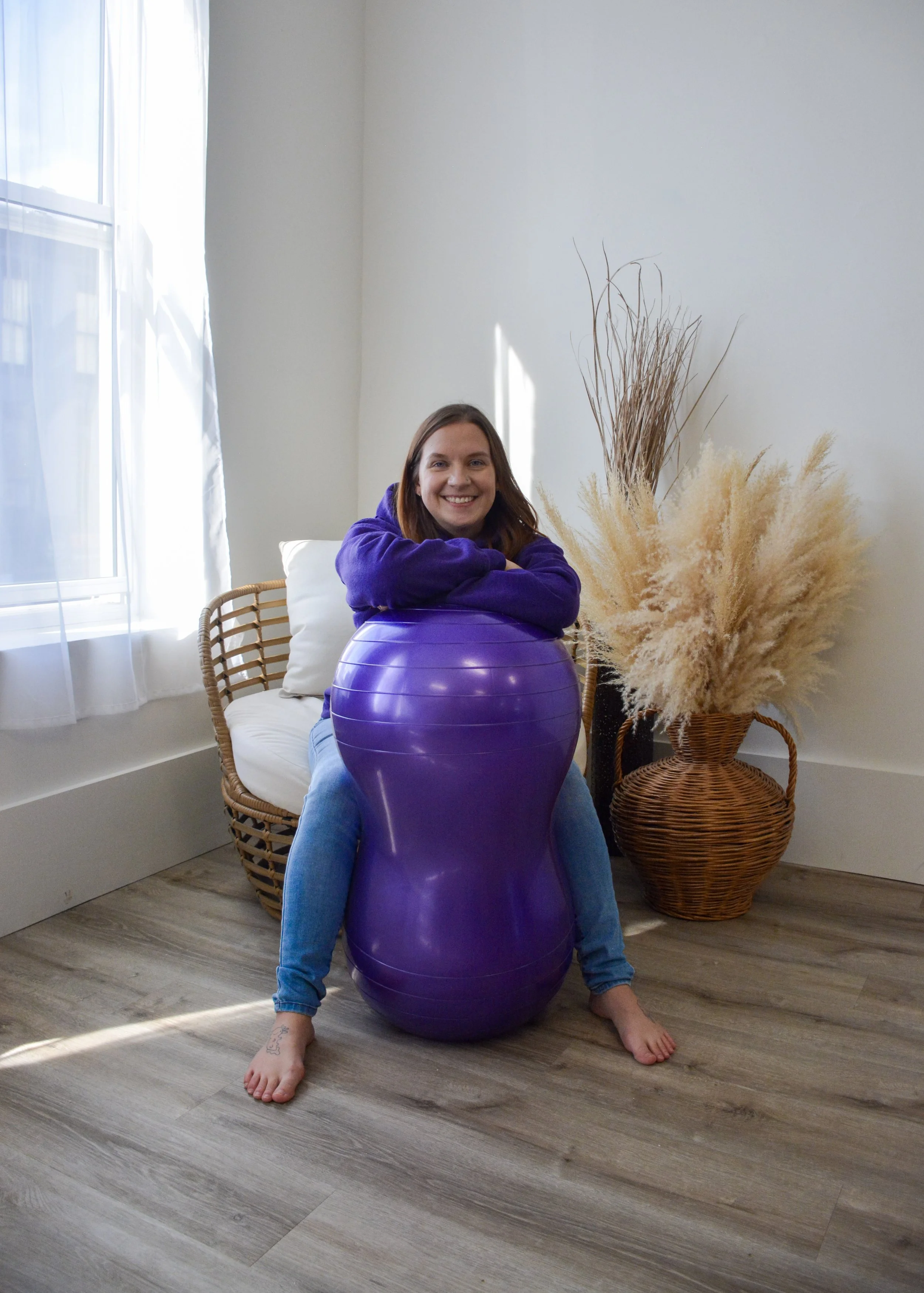 A woman sitting on a purple peanut ball, smiling, with her arms resting on the ball in a bright room with a wicker chair, white curtains, and a large vase of dried pampas grass.