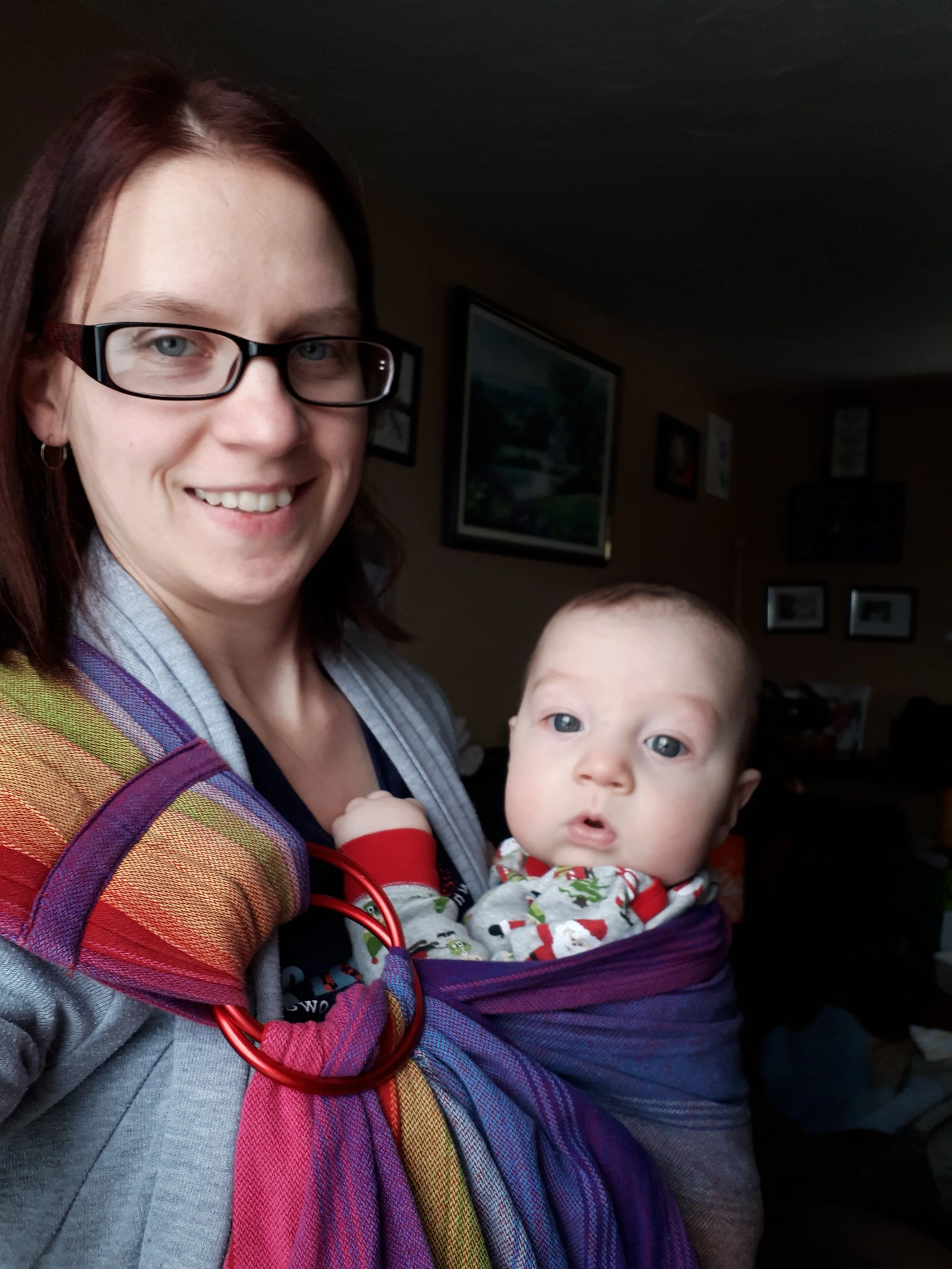 A woman with glasses smiling while holding a baby in a colorful wrap carrier indoors.
