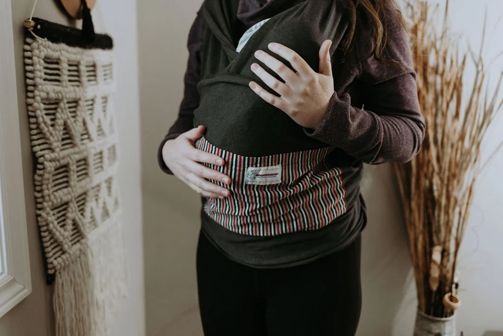 Person holding a baby in a gray baby carrier inside home with decorative dried plants.