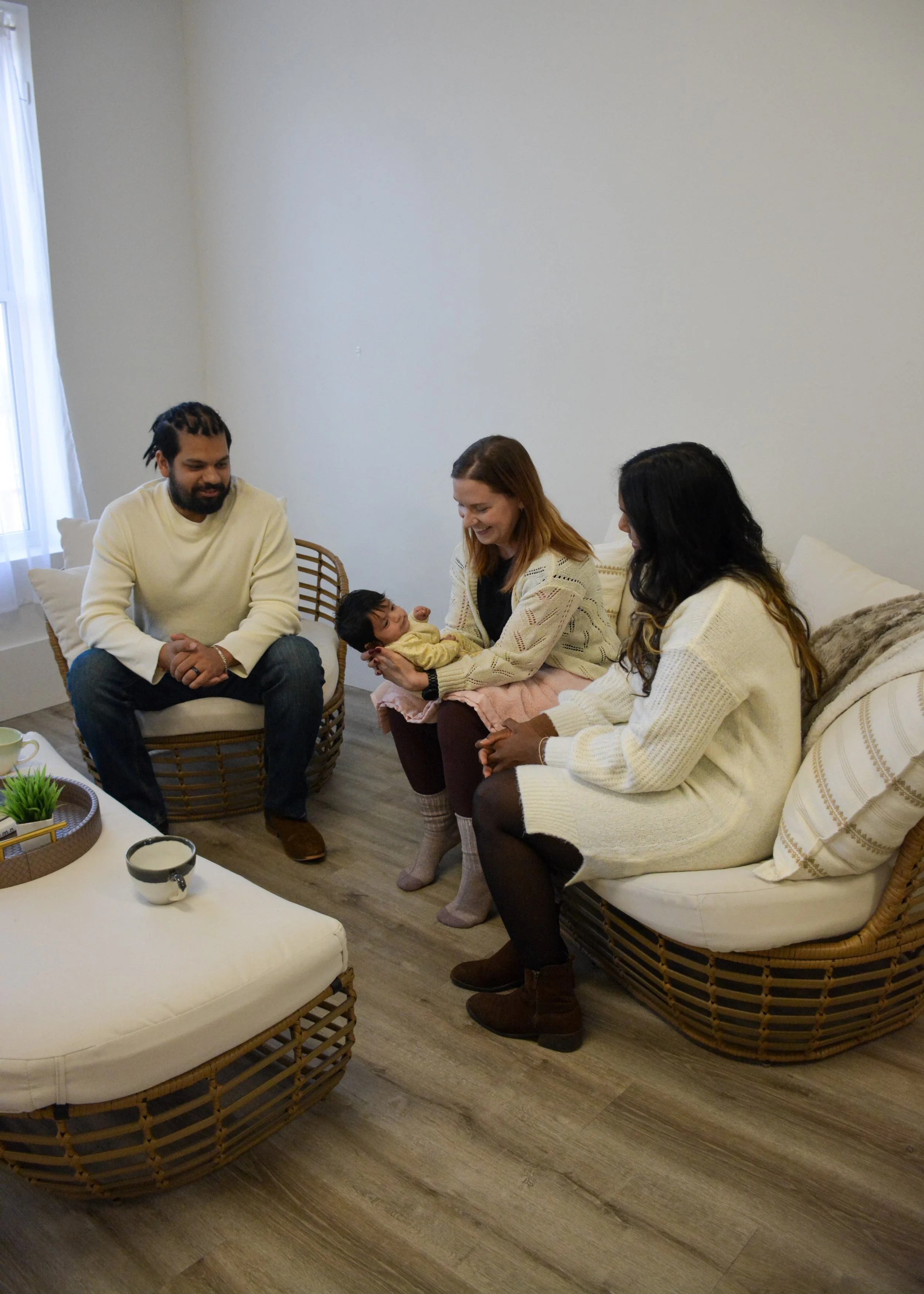 Group of three adults sitting in a living room, smiling and interacting with a baby held by a woman. The room has wicker furniture, a coffee table with plants and a cup, and a window with white curtains.