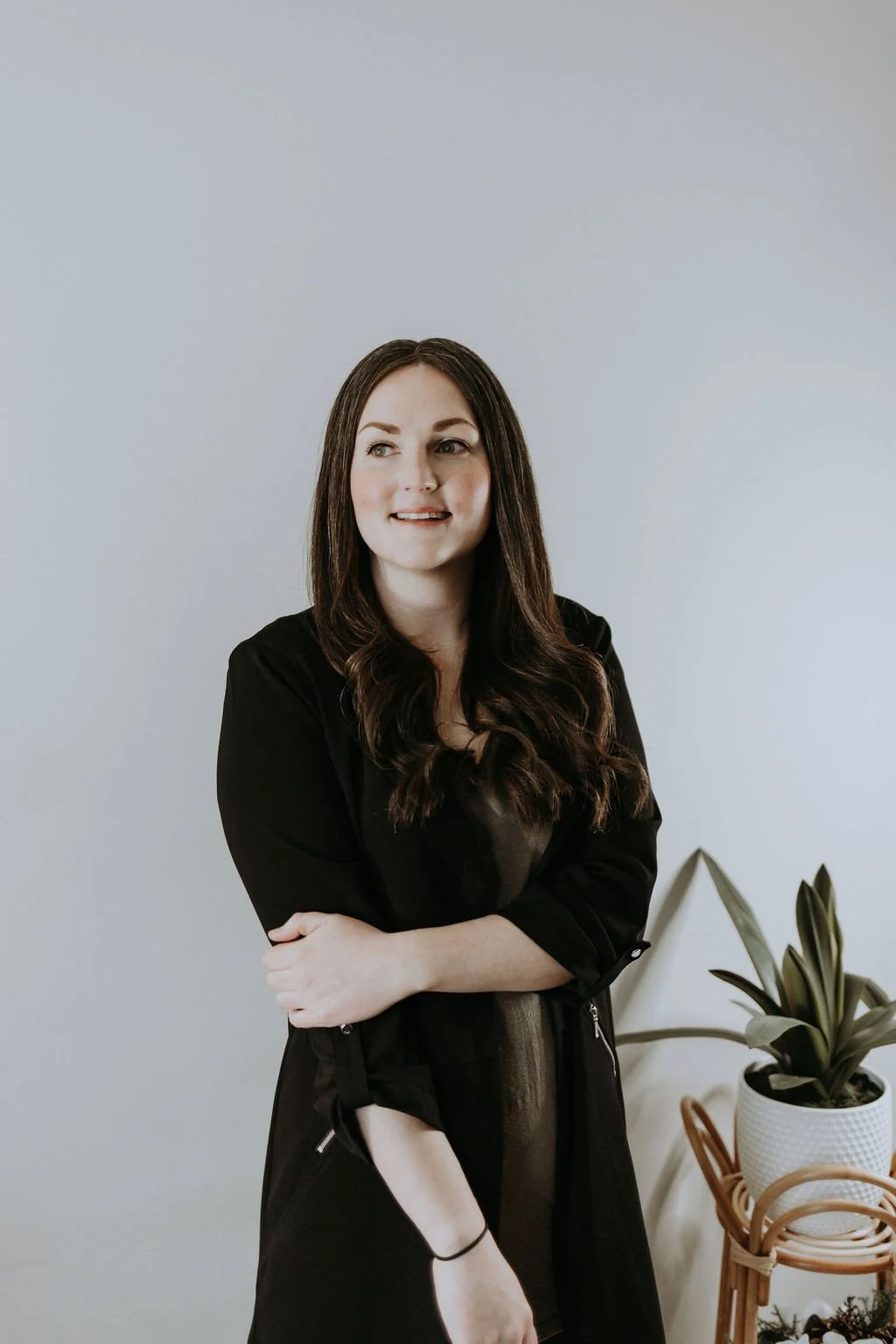 A woman with brown, wavy hair and light skin wearing a black shirt over a brown dress, standing indoors against a plain light-colored wall, with potted plants nearby.