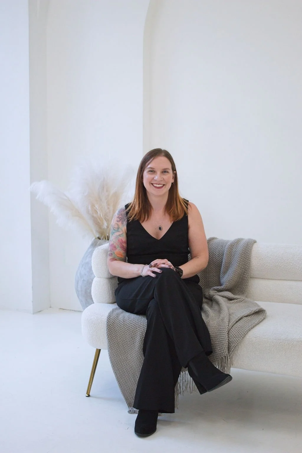 A smiling woman with brown hair and tattoos on her left arm, wearing a black sleeveless top and black pants, sitting on a light-colored sofa with a blanket, in a bright, minimalist room with white walls, and a large vase with pampas grass behind her.