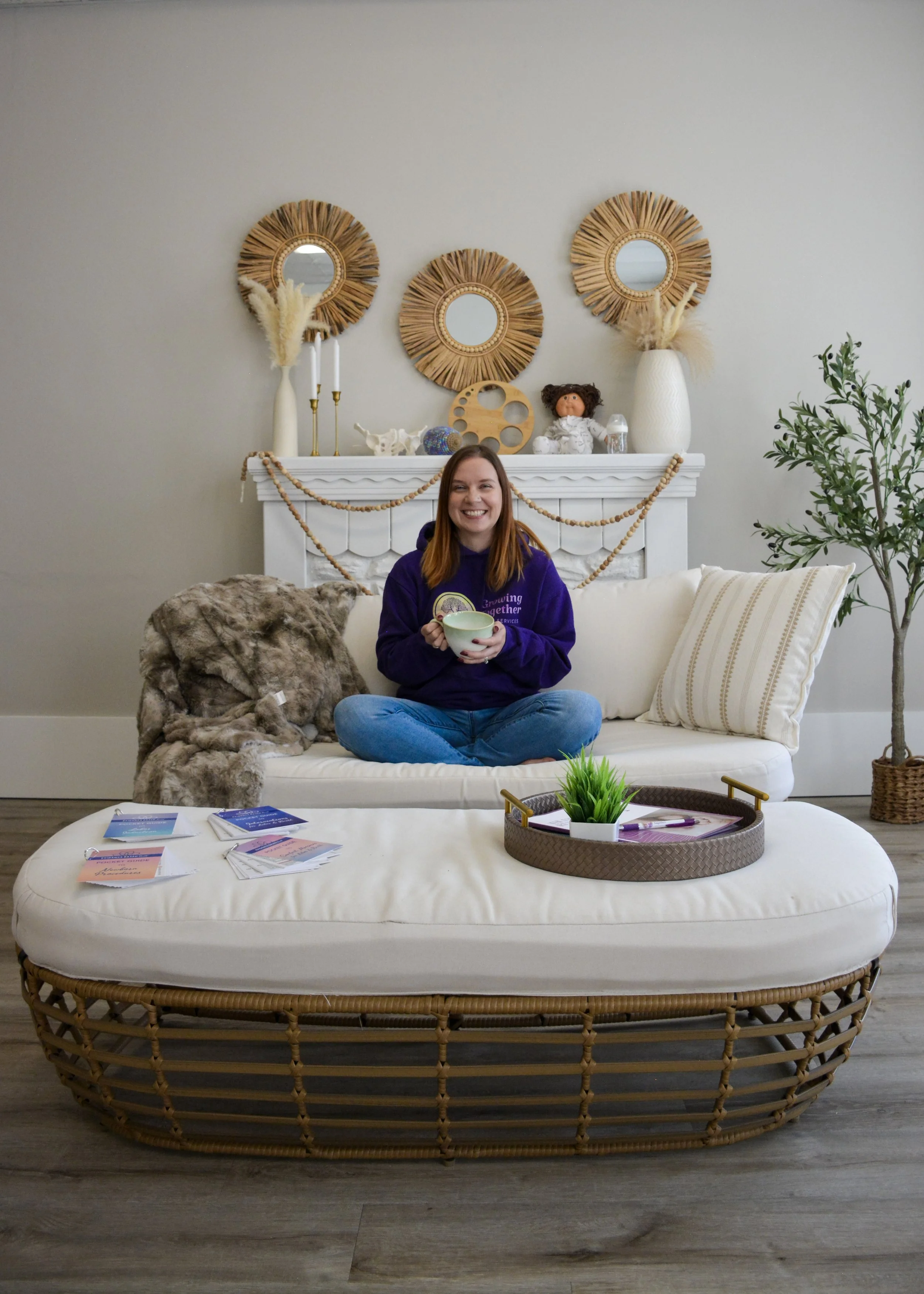 A woman sitting cross-legged on a white couch, holding a mug, smiling in a cozy living room with beige walls, decorative mirrors, plants, and books.