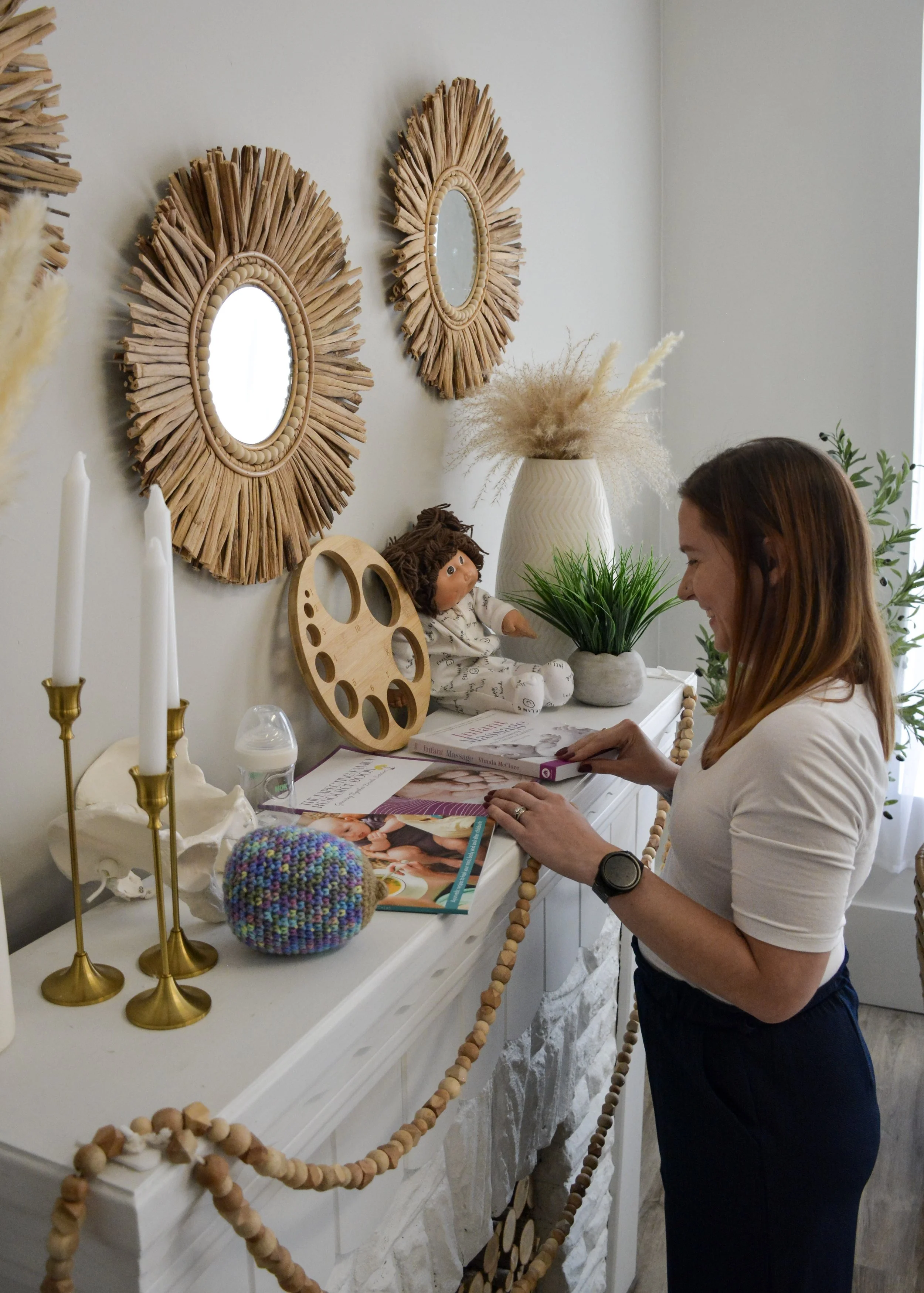 A woman with shoulder-length brown hair, dressed in a white short-sleeve shirt and dark pants, is decorating a white mantel with books, decorative items, and plants in a well-lit room with two artistically designed sunburst mirrors on the wall.
