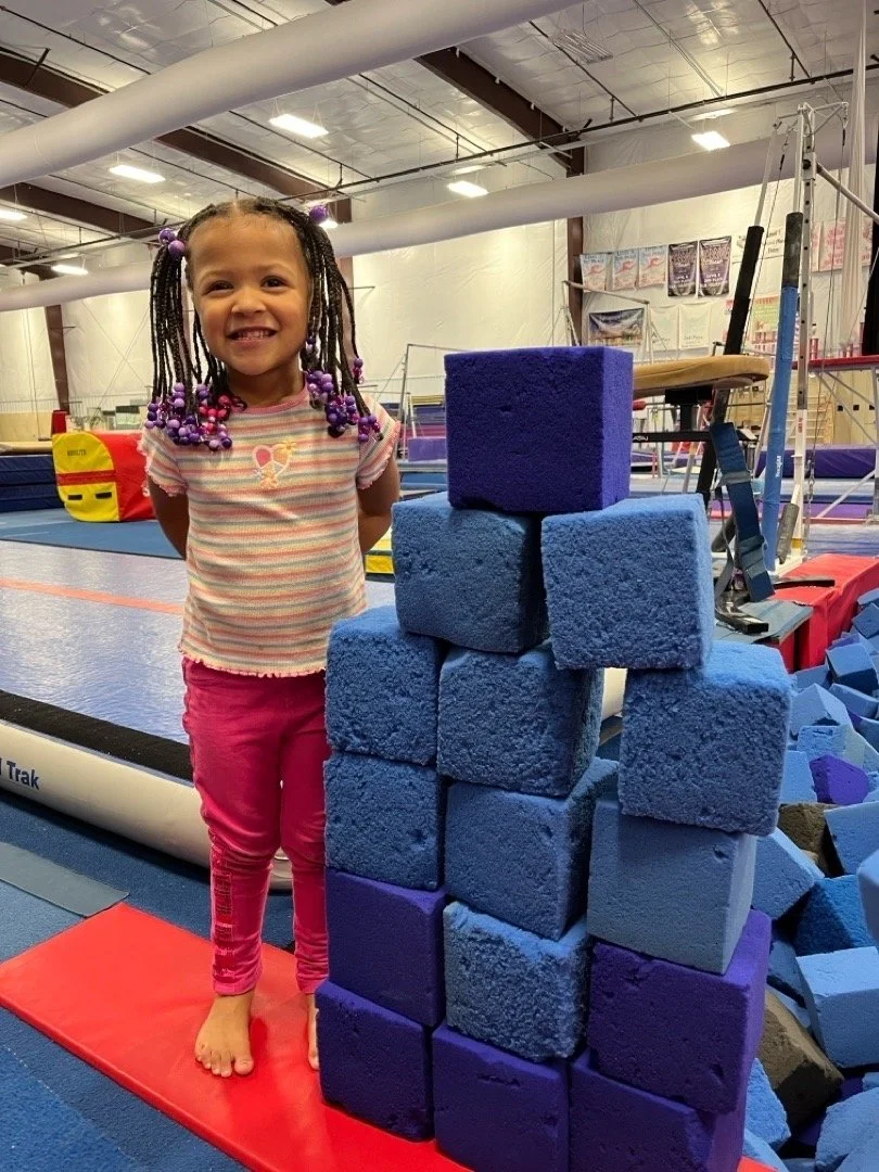 Smiling young girl with braided hair decorated with purple beads, standing barefoot on a red gymnastics mat in a gymnasium, next to a tall stack of blue and purple foam blocks.