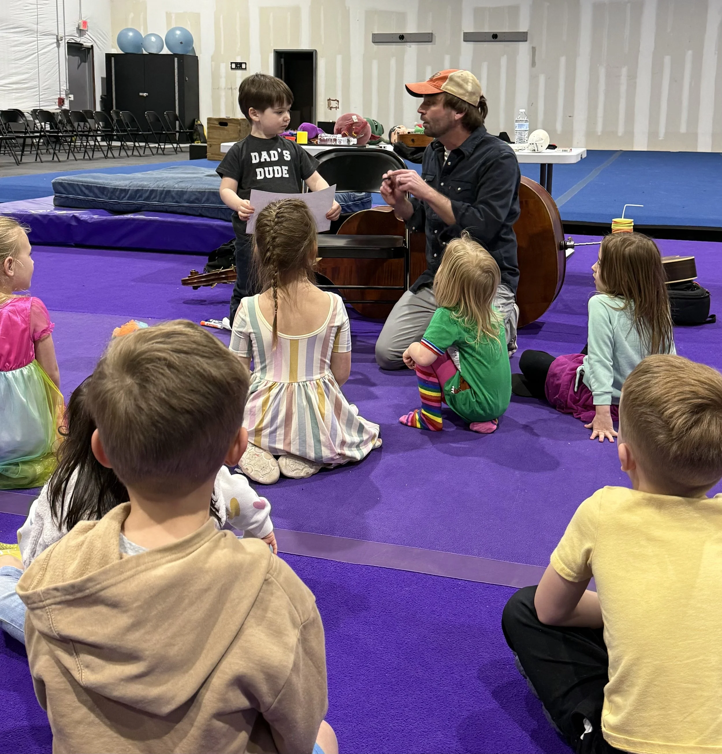 A man with long hair and a beard, wearing a cap and dark clothing, is kneeling and talking to a group of young children sitting on a purple mat. The children are listening attentively. There is a boy standing with a paper in his hand, and the background has gym equipment and mats.