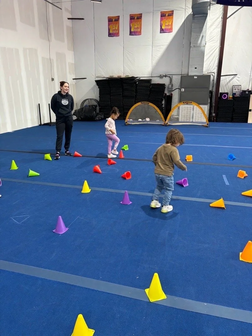 Two young children and an adult in an indoor gym with blue flooring, playing with colorful cones set up as an obstacle course.