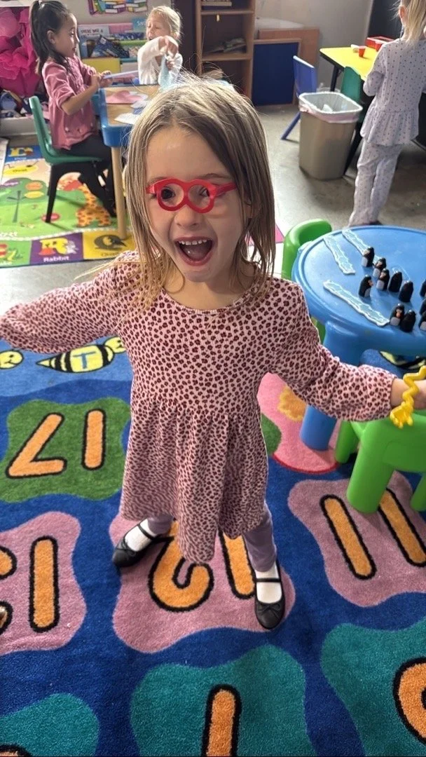 A young girl with glasses, smiling and celebrating, standing on a colorful alphabet rug in a classroom with other children and classroom furniture in the background.