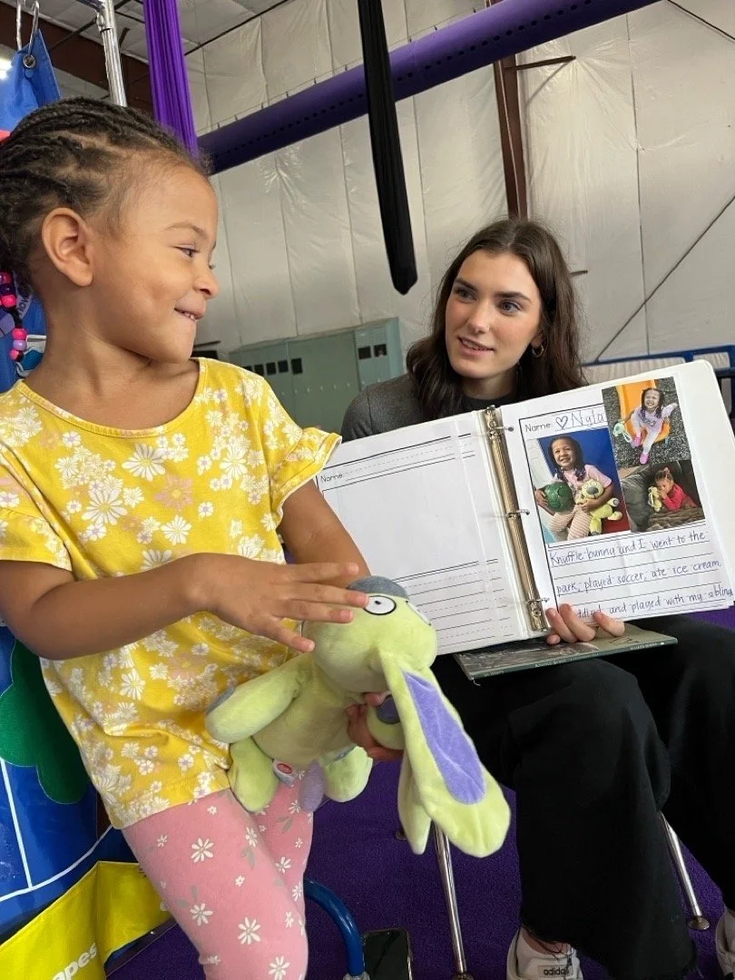 A young girl with braids and a yellow shirt with white flowers sits and plays with a stuffed animal, while an older girl with long dark hair and a gray shirt shows her a photo album with pictures of the girl holding a ball and a girl in a pink sweater, smiling at each other inside a gym.