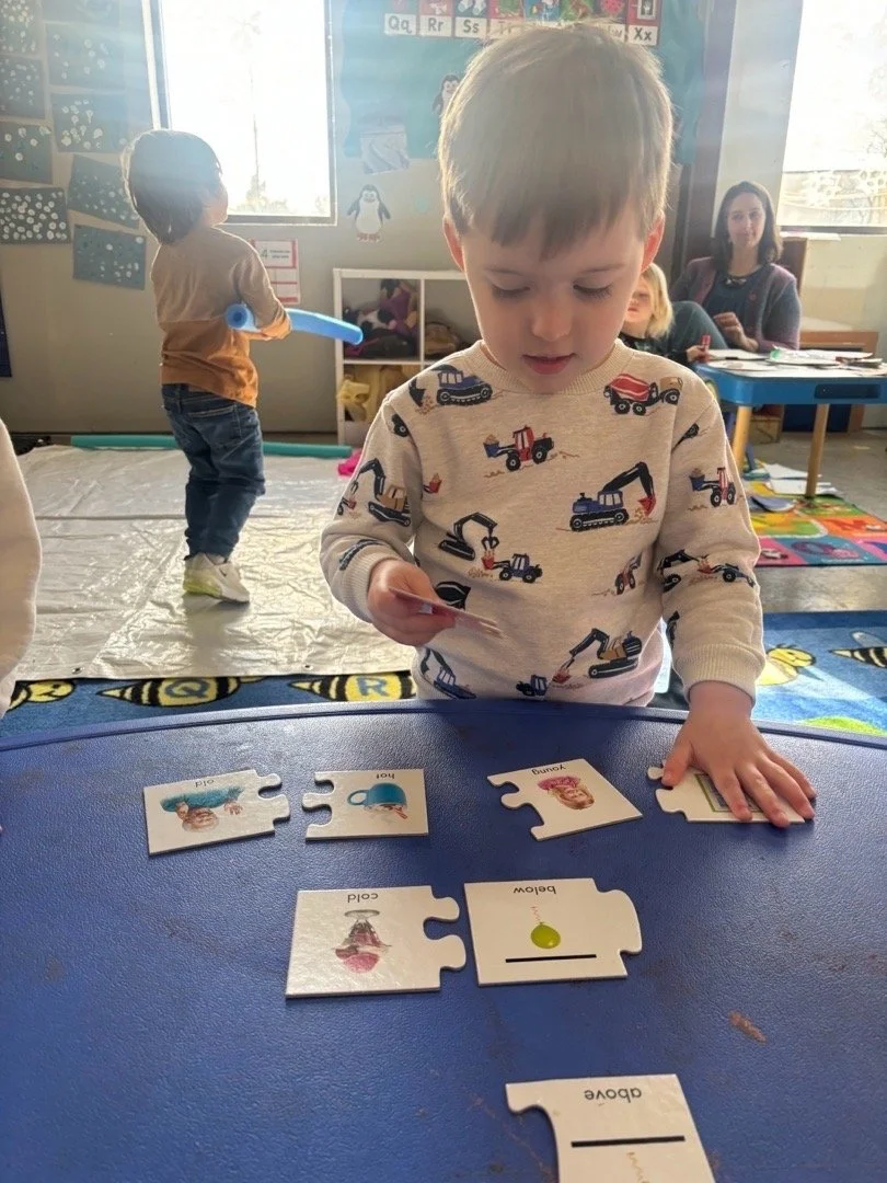 Child in a patterned shirt plays with puzzle pieces on a blue table, with another child in the background playing with a balloon in a colorful classroom.